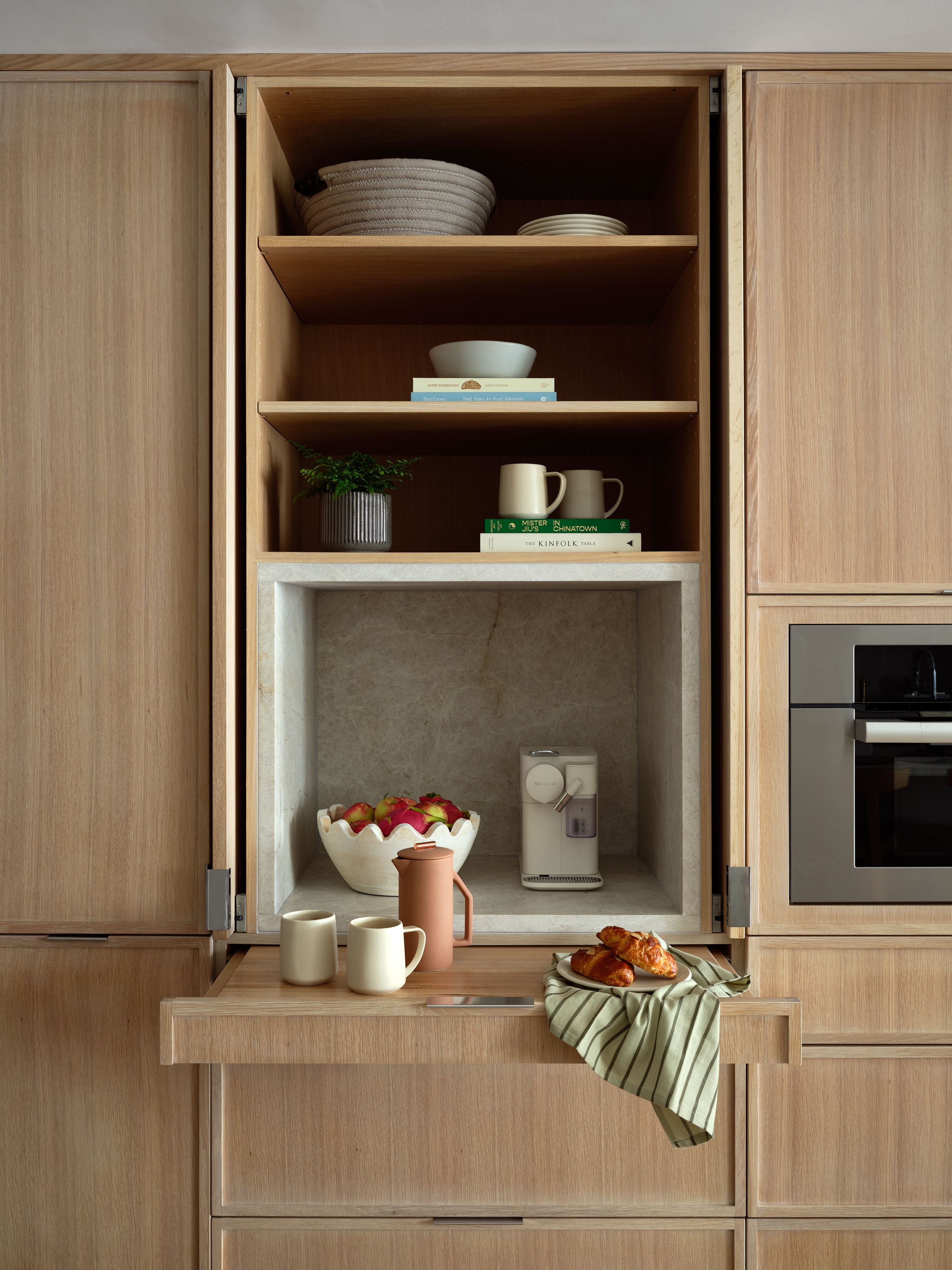 An open pantry cabinet with built-in oak shelving, dishware, and a marble-lined nook holding an espresso machine and pastries.