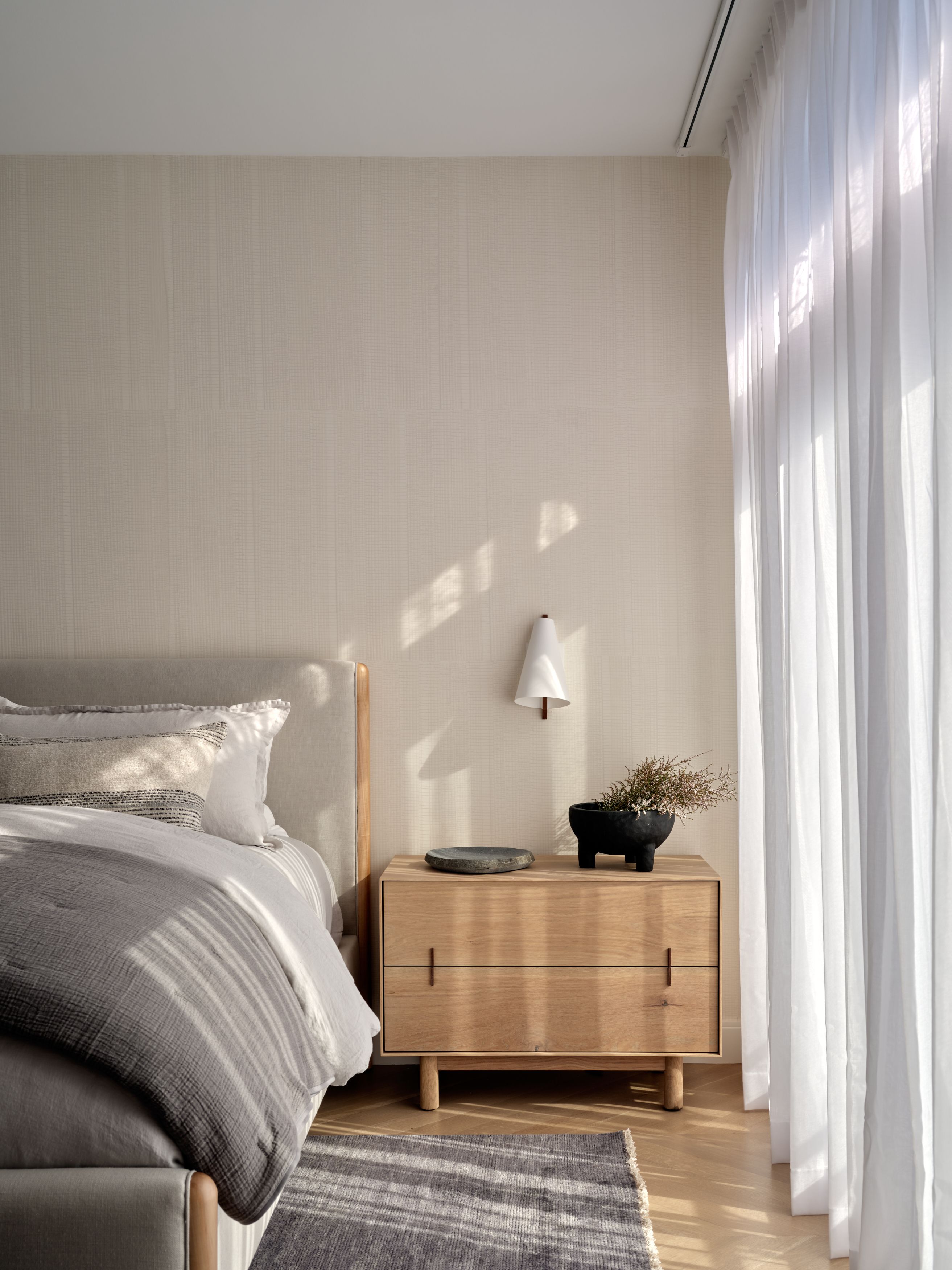 A bedroom nightstand corner with a cone-shaped wall sconce, grasscloth wallpaper, a white oak nightstand, and sheer curtains.