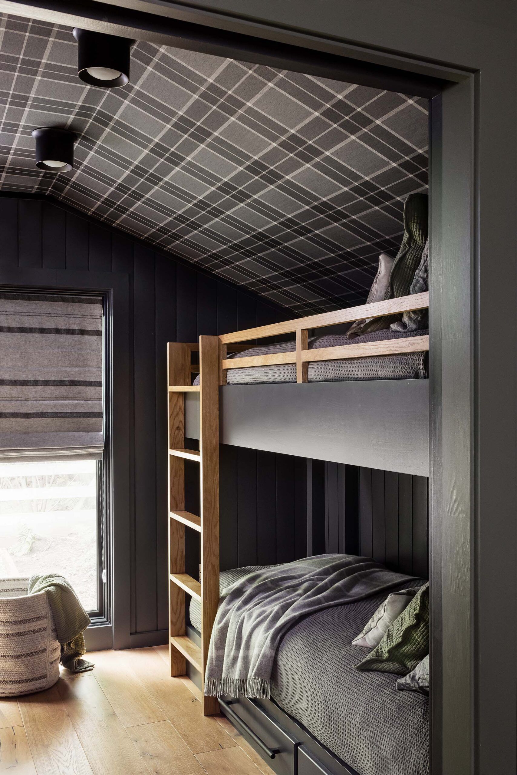 Built-in bunk beds with a plaid-papered ceiling, dark gray cabinetry, a wood ladder, and a striped roman shade.