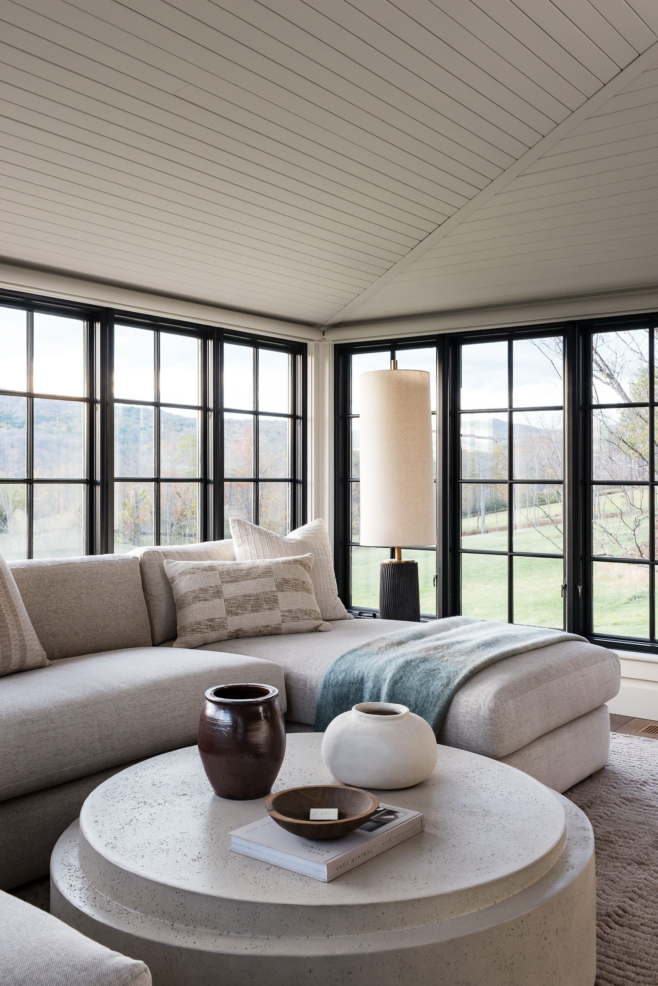 A sunroom corner with a bouclé-gray sectional sofa and a round stone coffee table is wrapped in black-framed windows overlooking open fields.