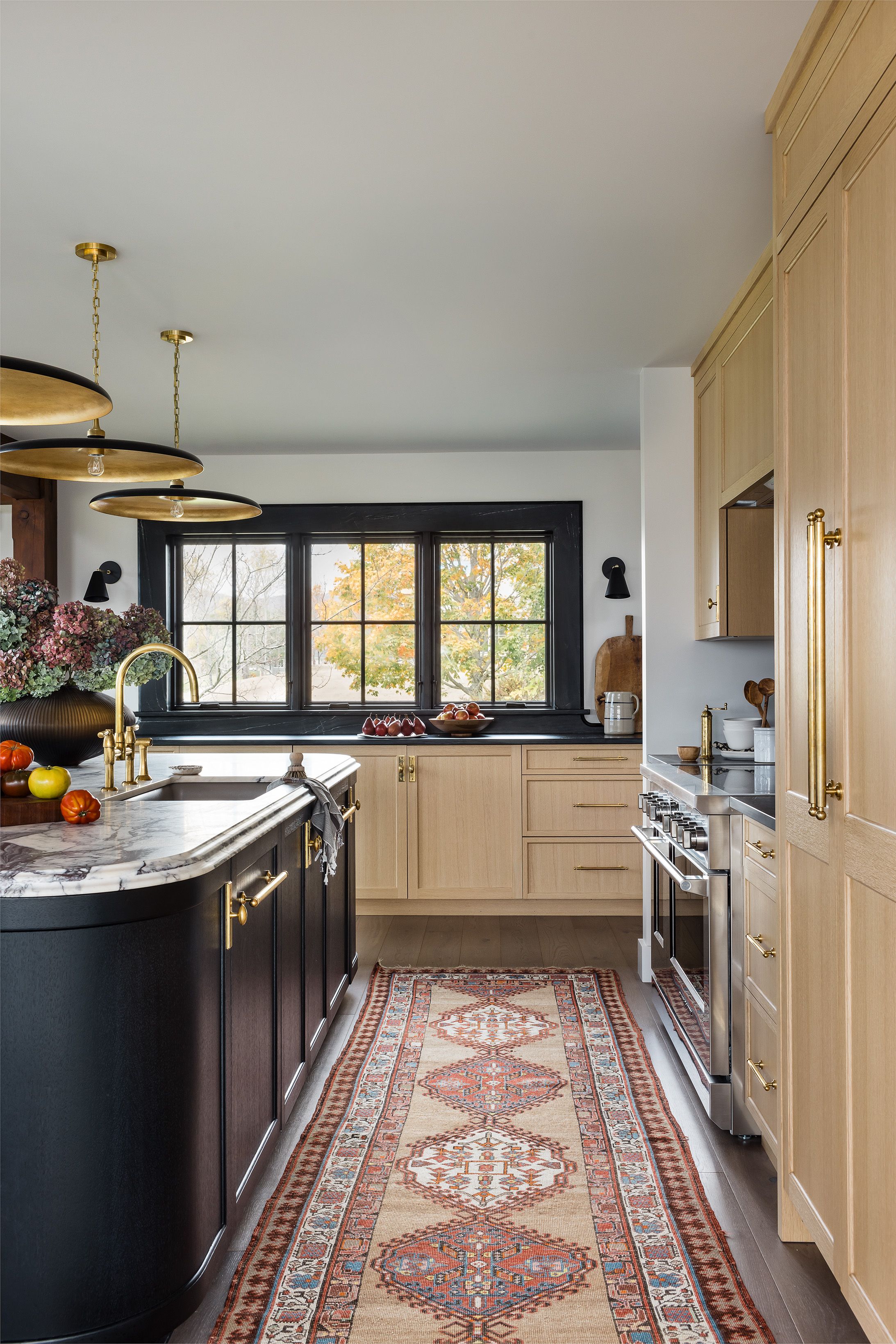 A kitchen with black cabinetry and marble counters has a black-trimmed window wall, brass disc pendants, and a vintage patterned runner rug.