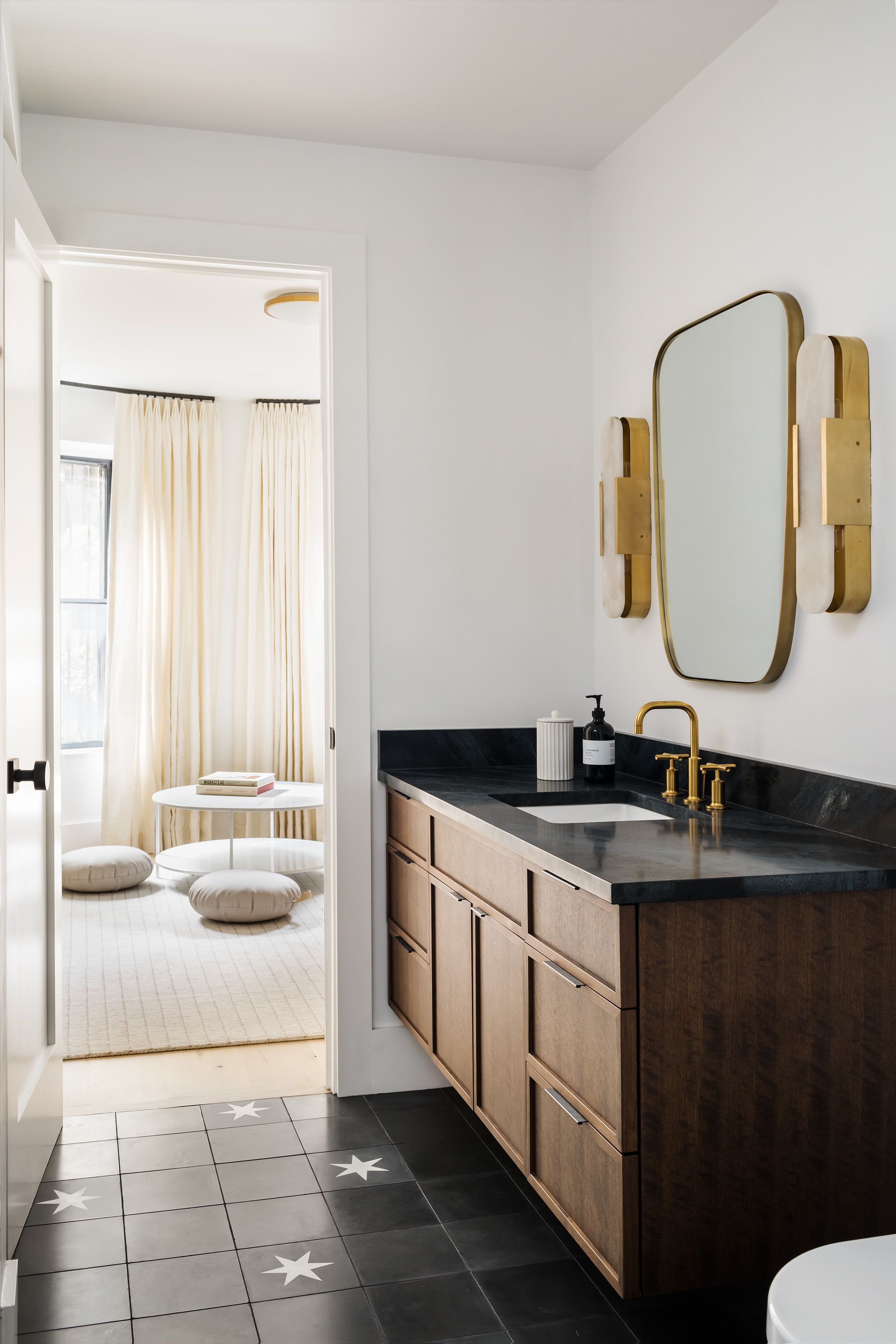 Bathroom with a round brass-framed mirror, brass sconces, and a black stone counter opens to a sitting room with floor cushions.