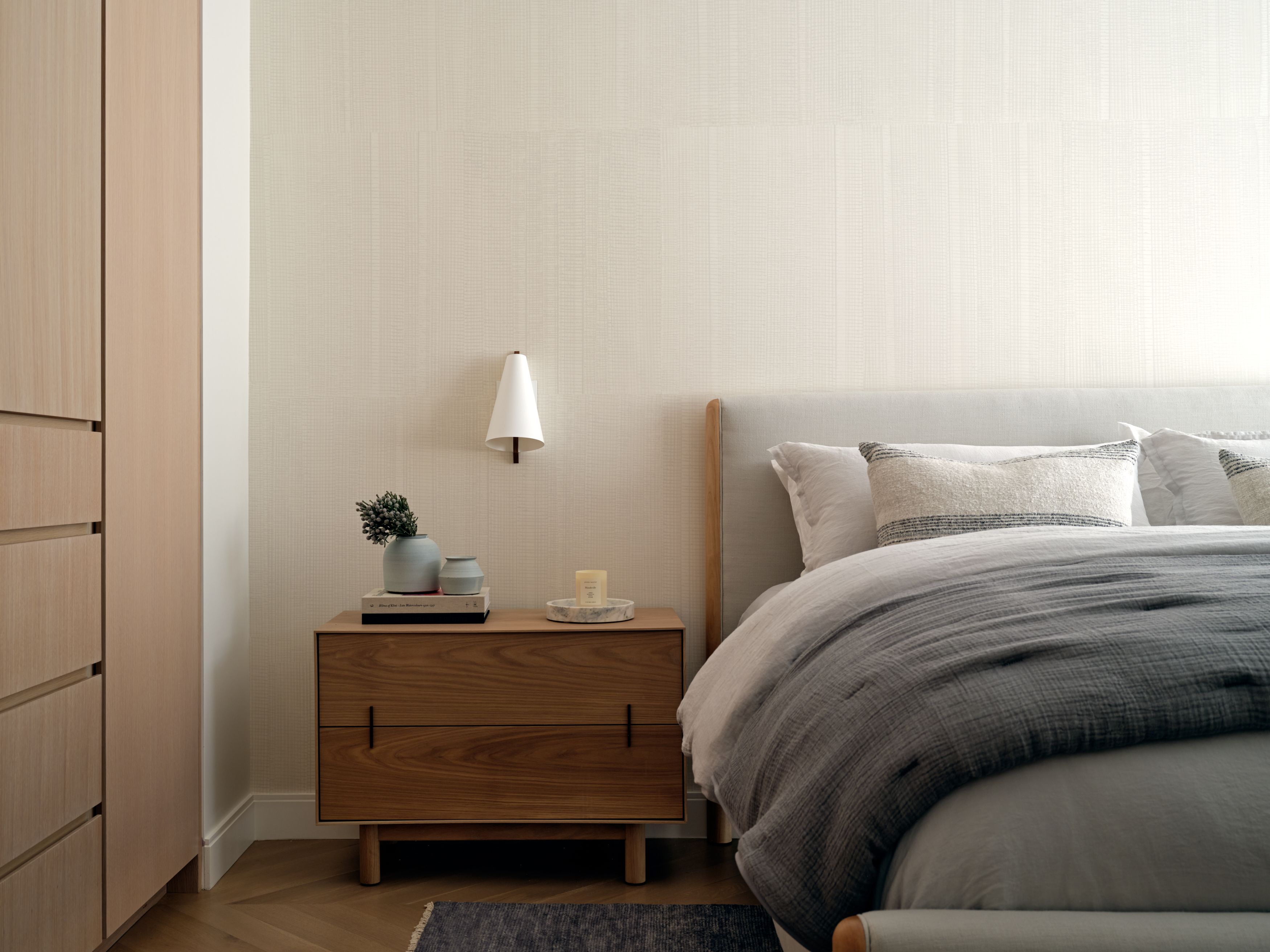 A bedroom with a walnut nightstand, a cone-shaped pendant sconce, an upholstered headboard, and a herringbone wood floor.