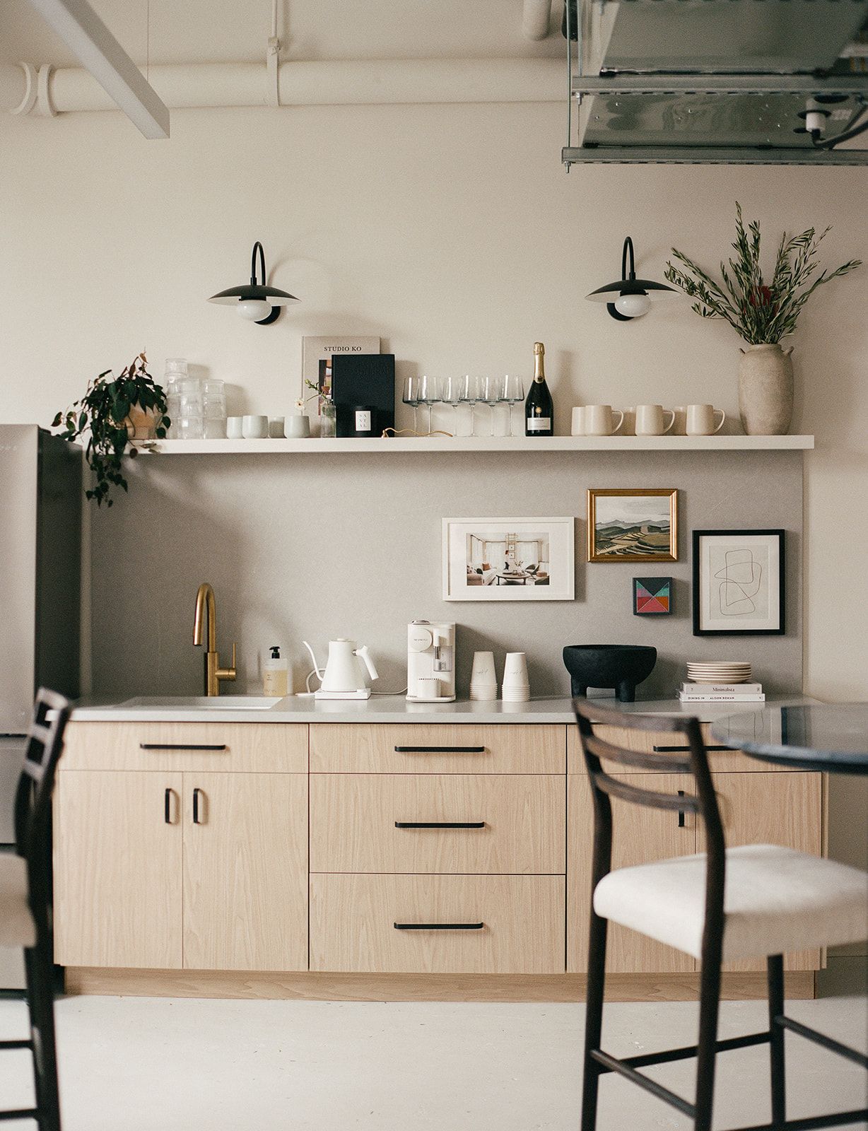 A studio kitchenette with black wall sconces, a gold faucet, an open shelf of glassware and champagne, and a gallery wall of framed art.