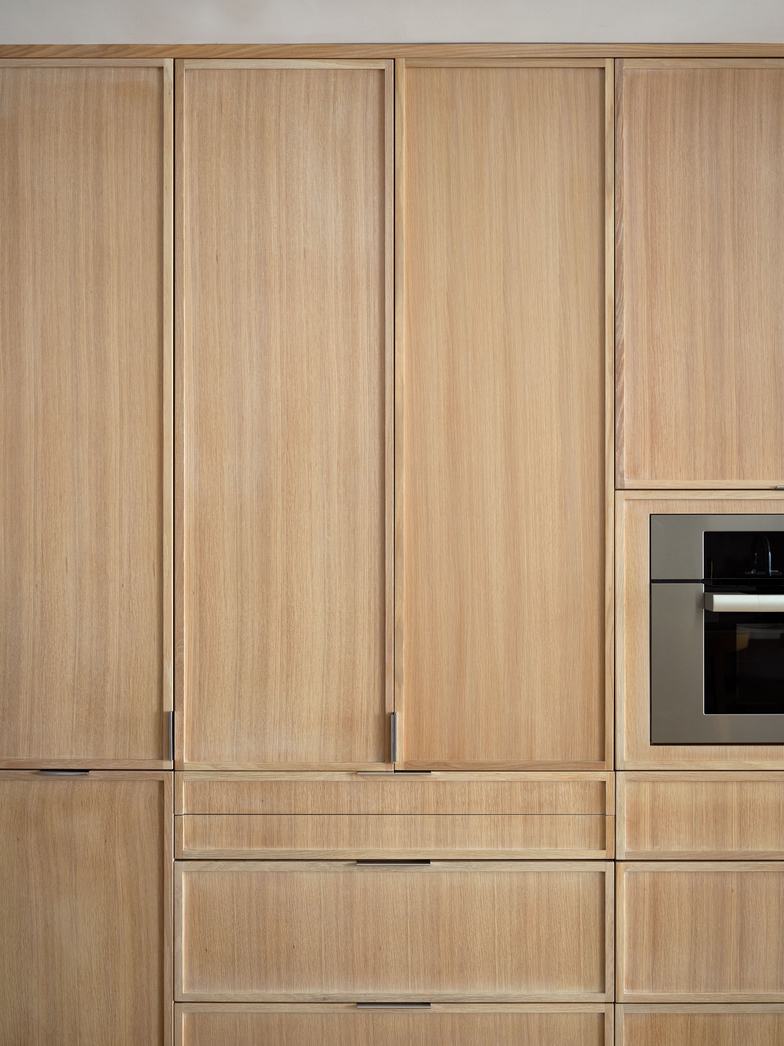 A close-up of vertical-grain white oak kitchen cabinetry with a built-in stainless steel oven and slim black pulls.