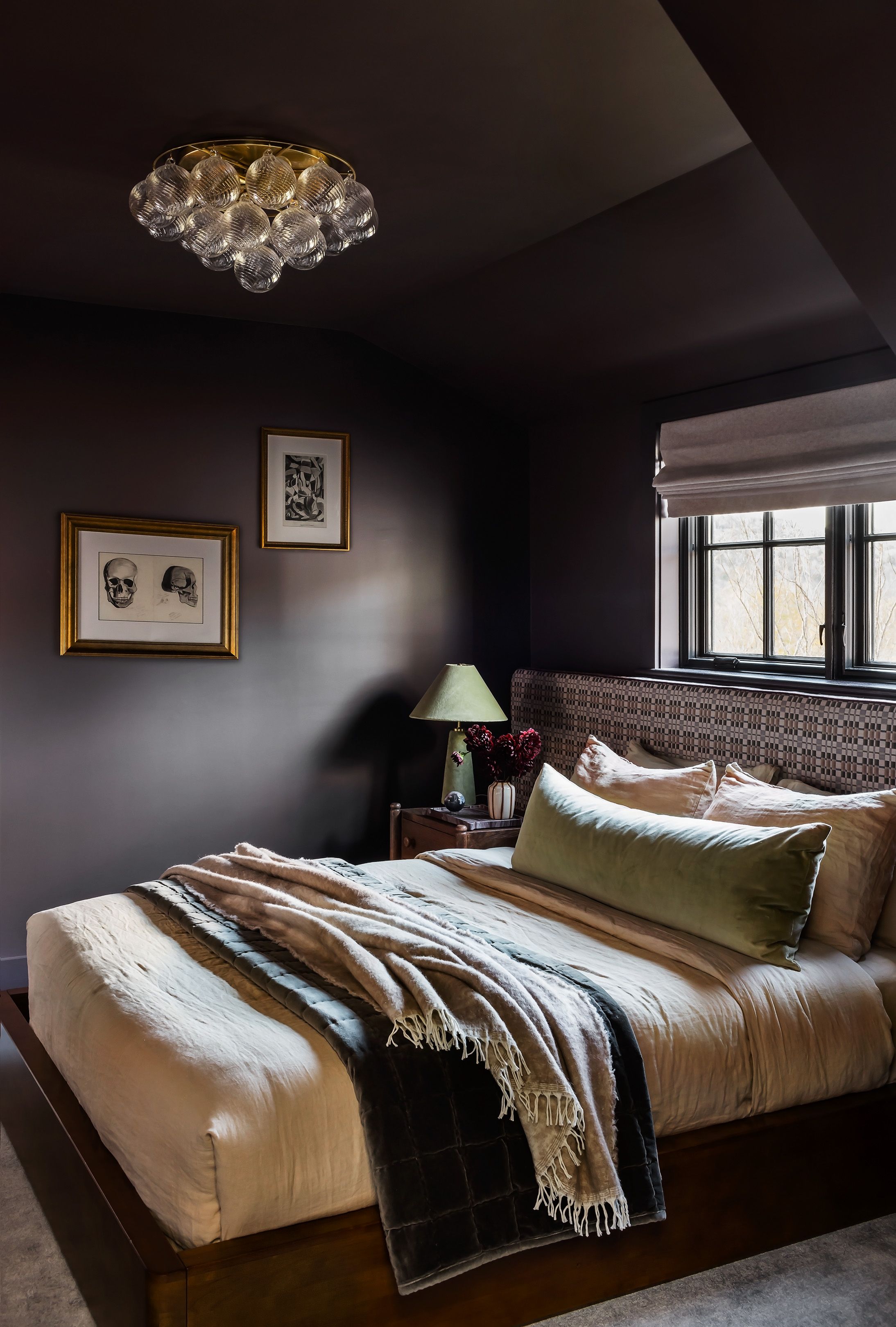 A dark plum bedroom pairs a checkered woven headboard with gold-framed anatomical skull prints and a bubbled glass flush-mount light.
