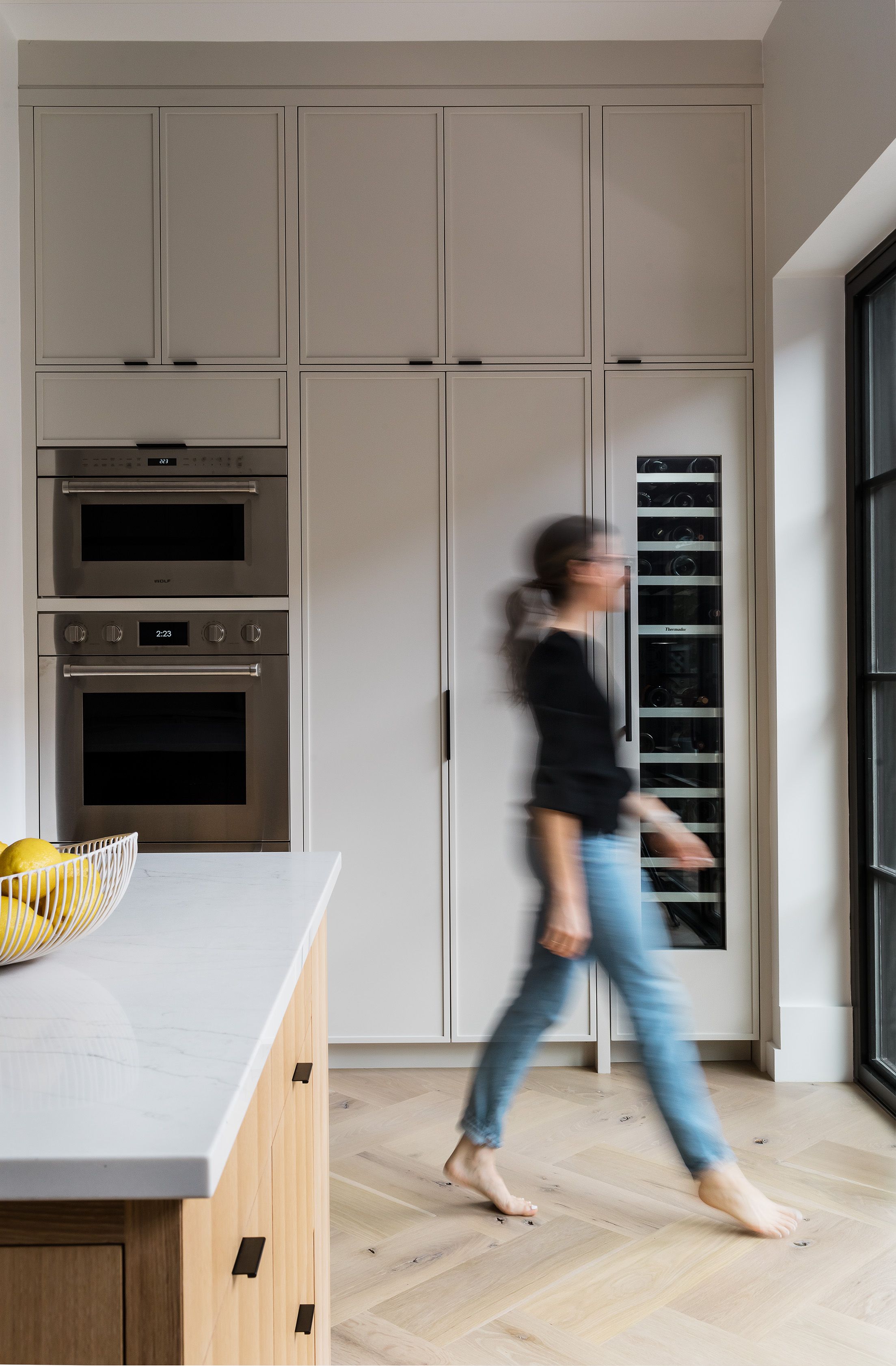 Someone walks barefoot through a kitchen with pale floor-to-ceiling cabinetry, a built-in wine fridge, and a marble island.