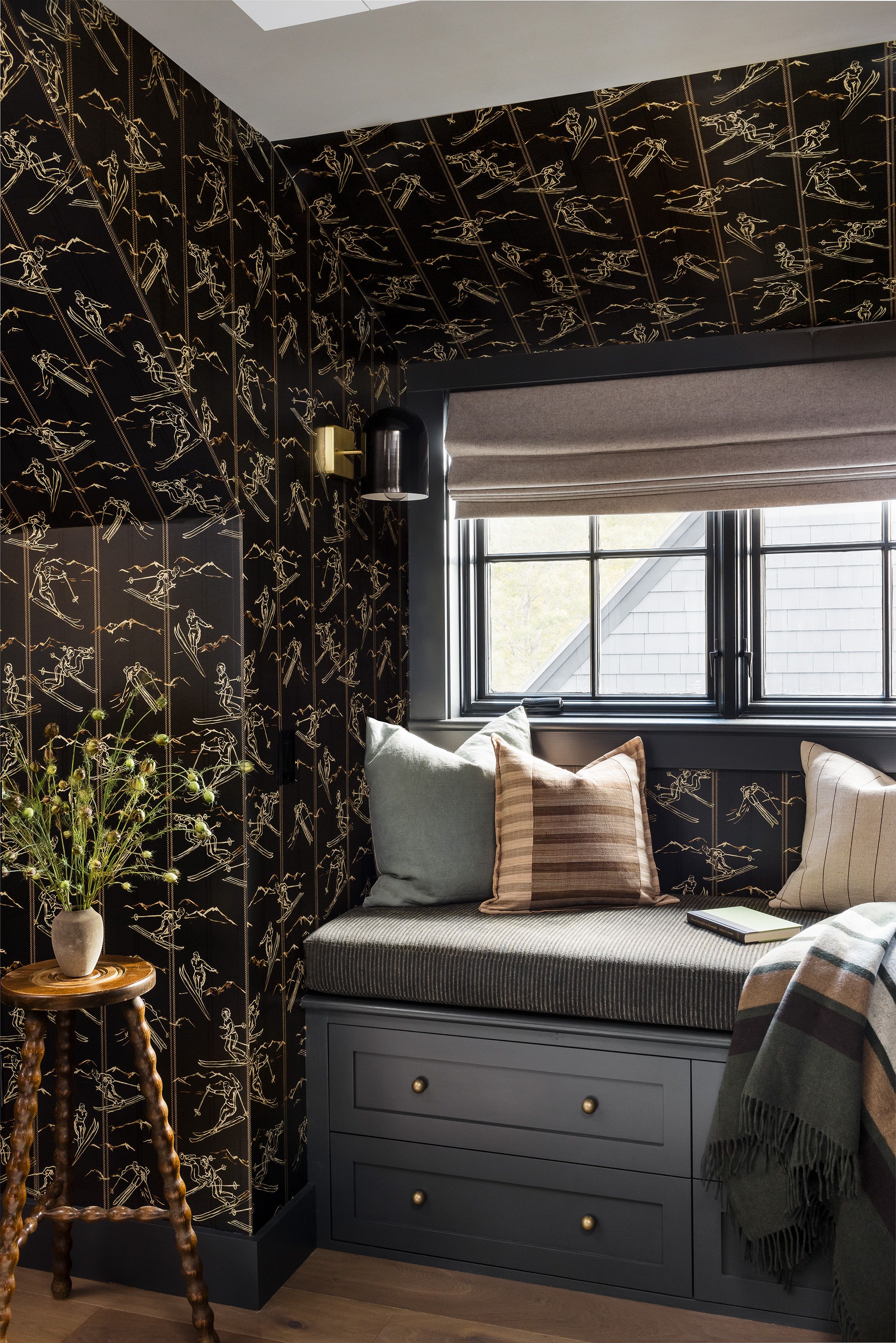 A window seat with striped and plaid pillows sits beneath gold ski-print wallpaper, above storage drawers beside a turned wood side table.