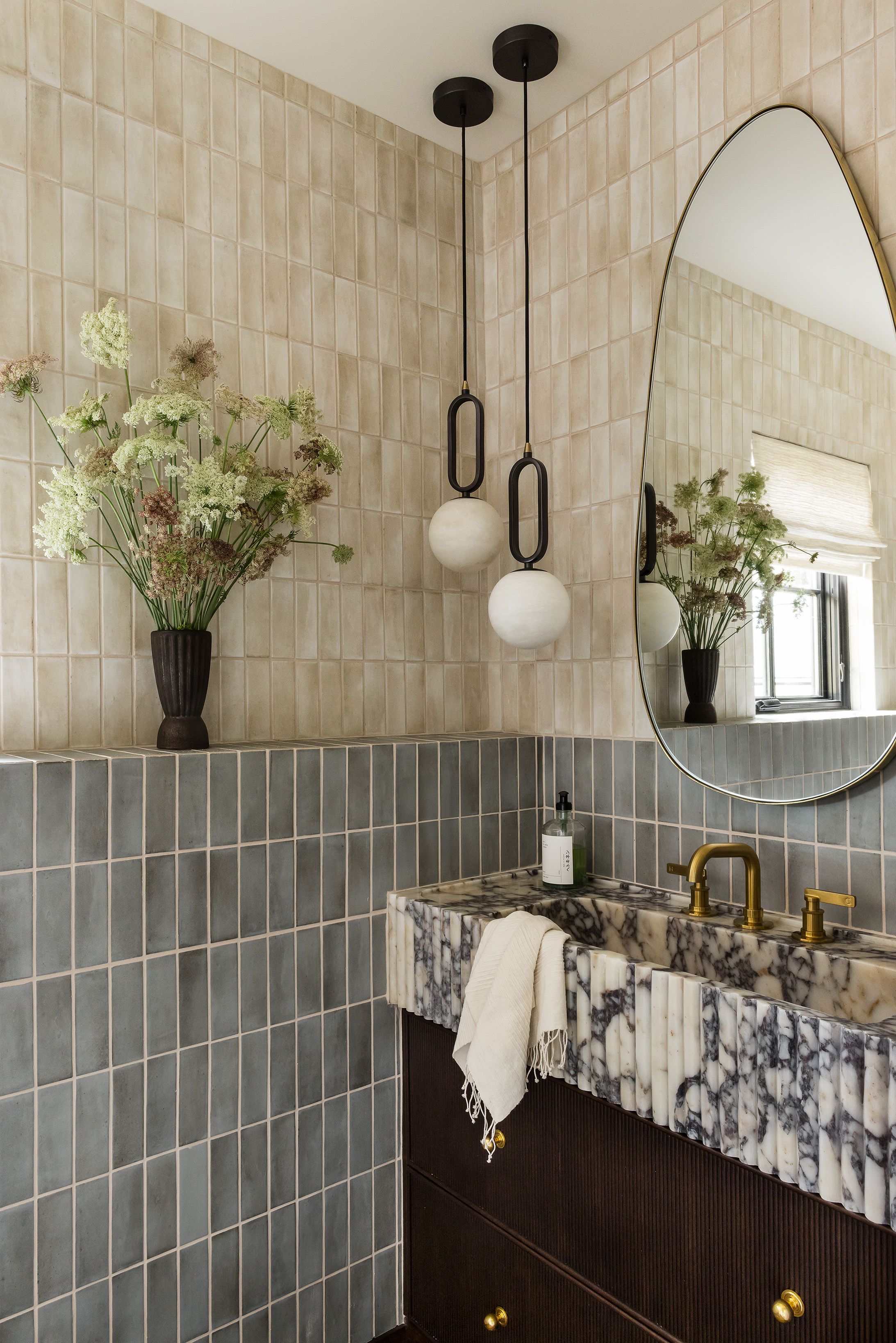 A bathroom with cream zellige-style tile above sage-green square tile pairs globe pendant lights with a fluted marble vanity and brass fixtures.