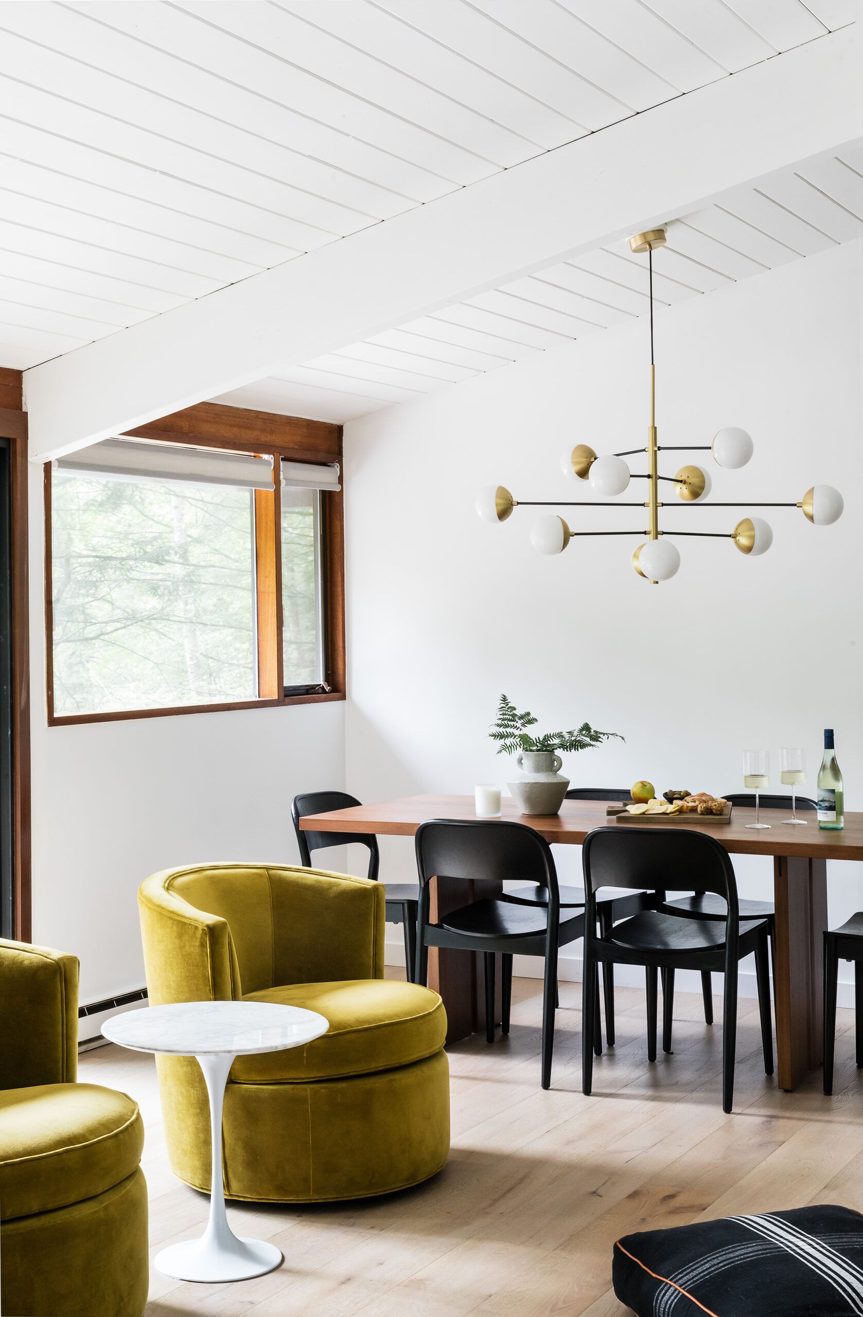 A dining area with a wood table, black modern chairs, a brass multi-globe chandelier, and a mustard velvet swivel chair nearby.