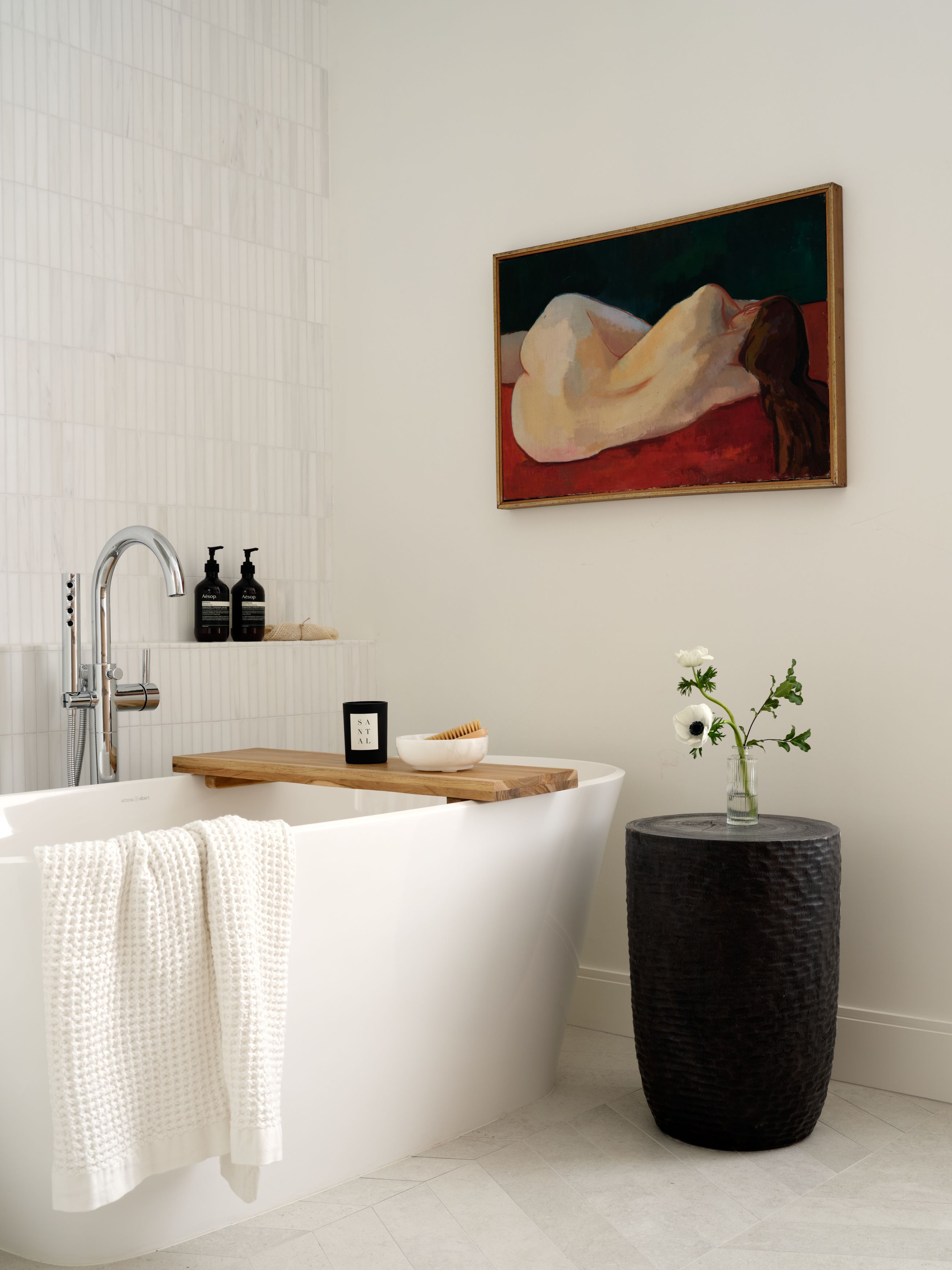 A primary bathroom with a painting of a reclining nude, a freestanding tub with a wood tray, and a carved black side table.