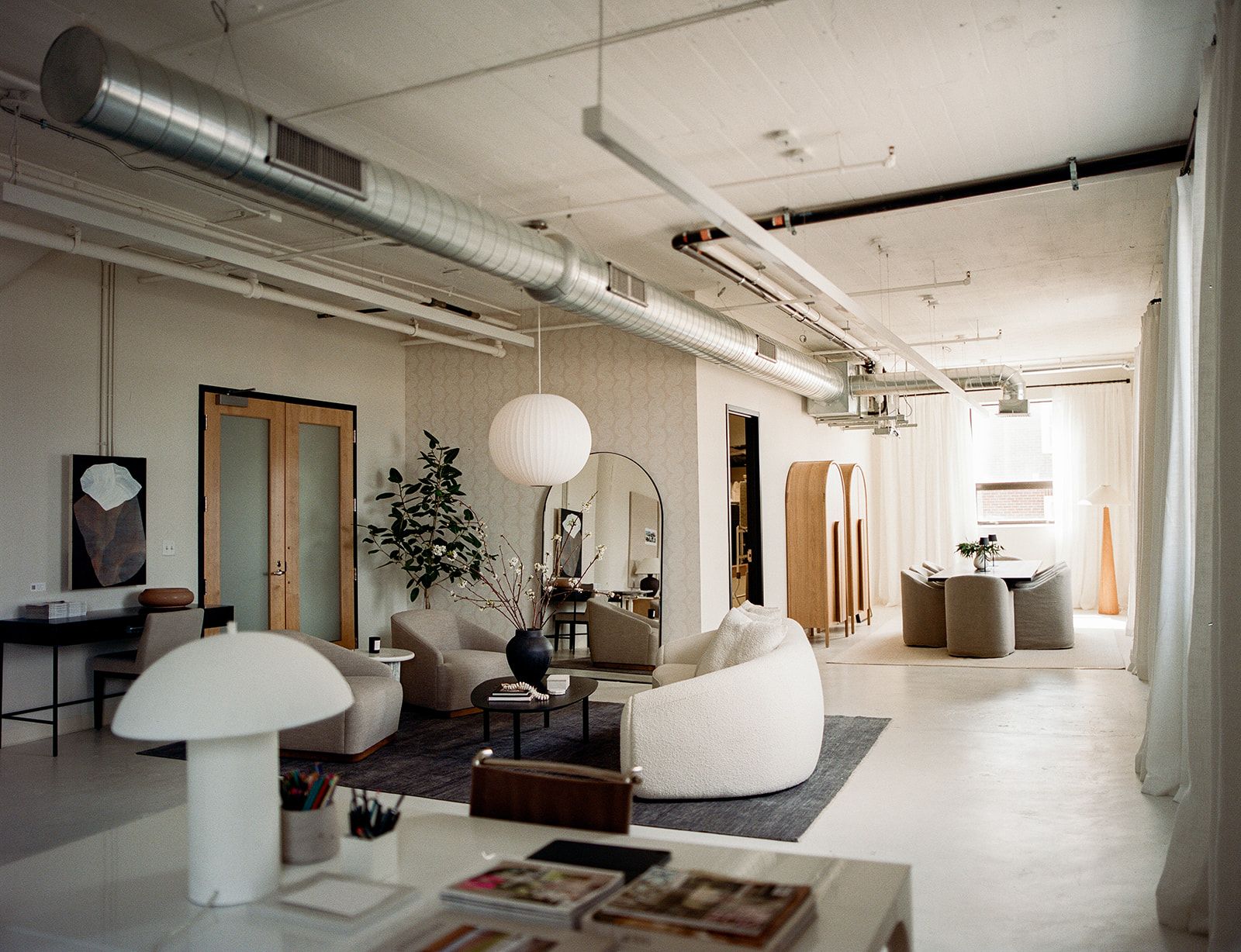 A loft space with exposed ductwork, round paper pendant lamps, cream boucle lounge chairs, and an arched floor mirror.