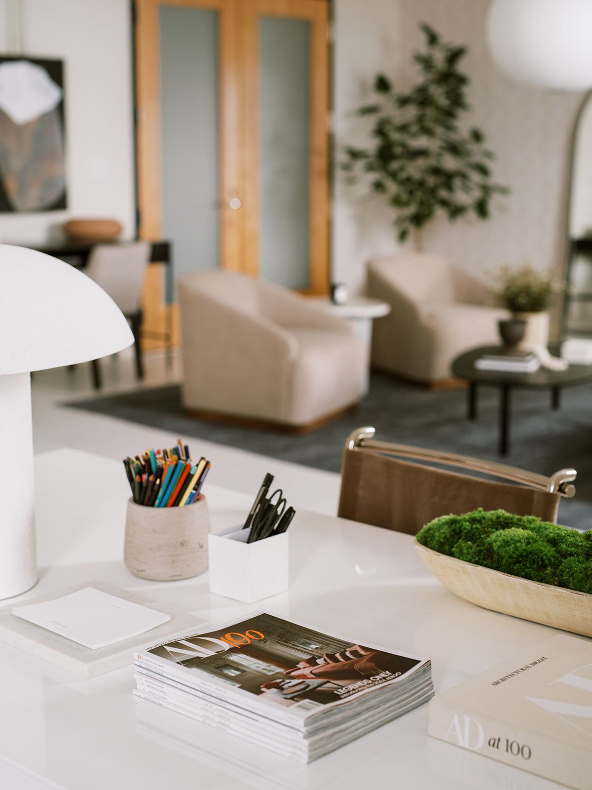 A studio desk with a white mushroom lamp, cups of pens and pencils, a bowl of moss, and lounge seating blurred in the background.