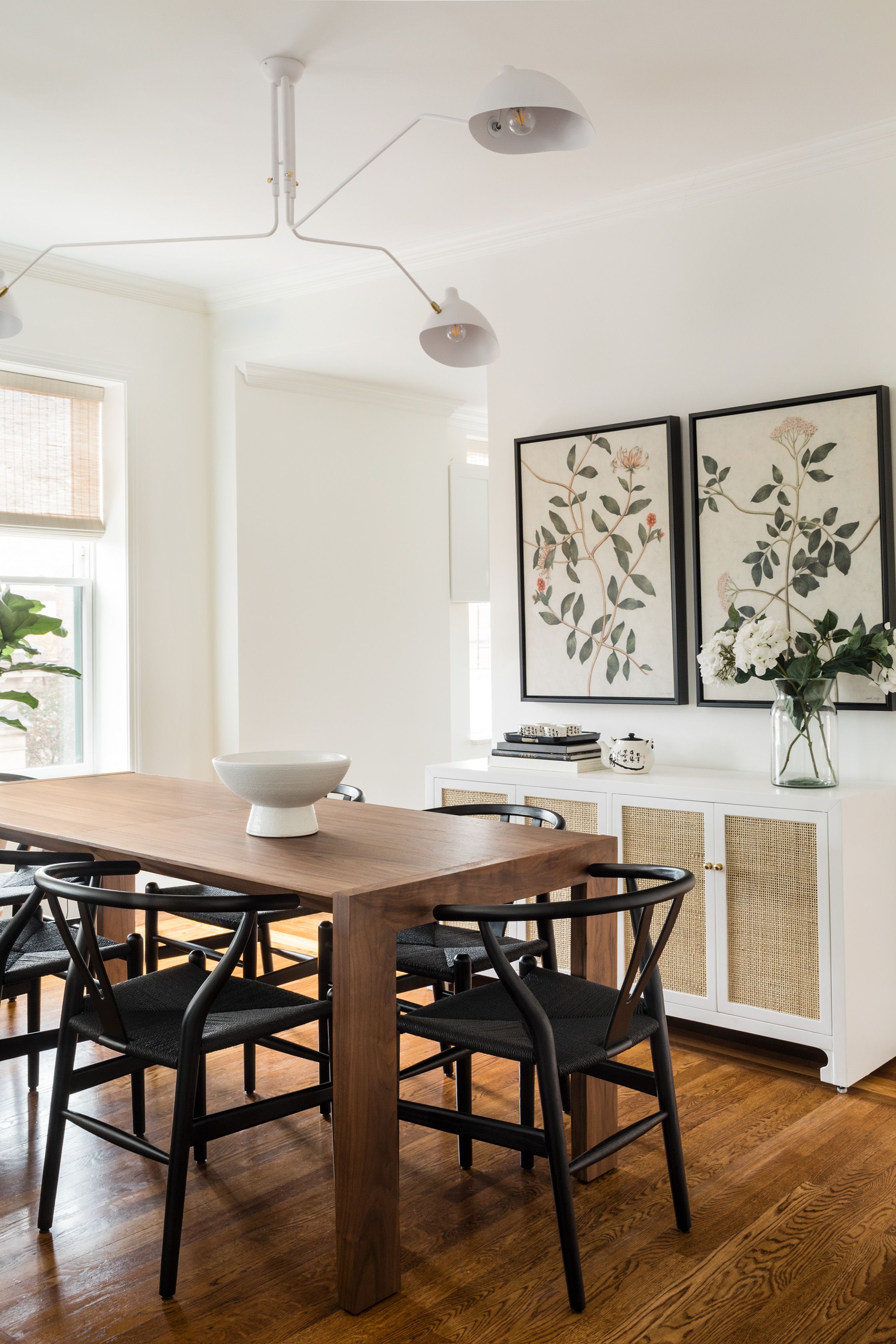 A dining room with a walnut table, black wishbone chairs, a cane-front sideboard, and a pair of framed botanical prints above it.