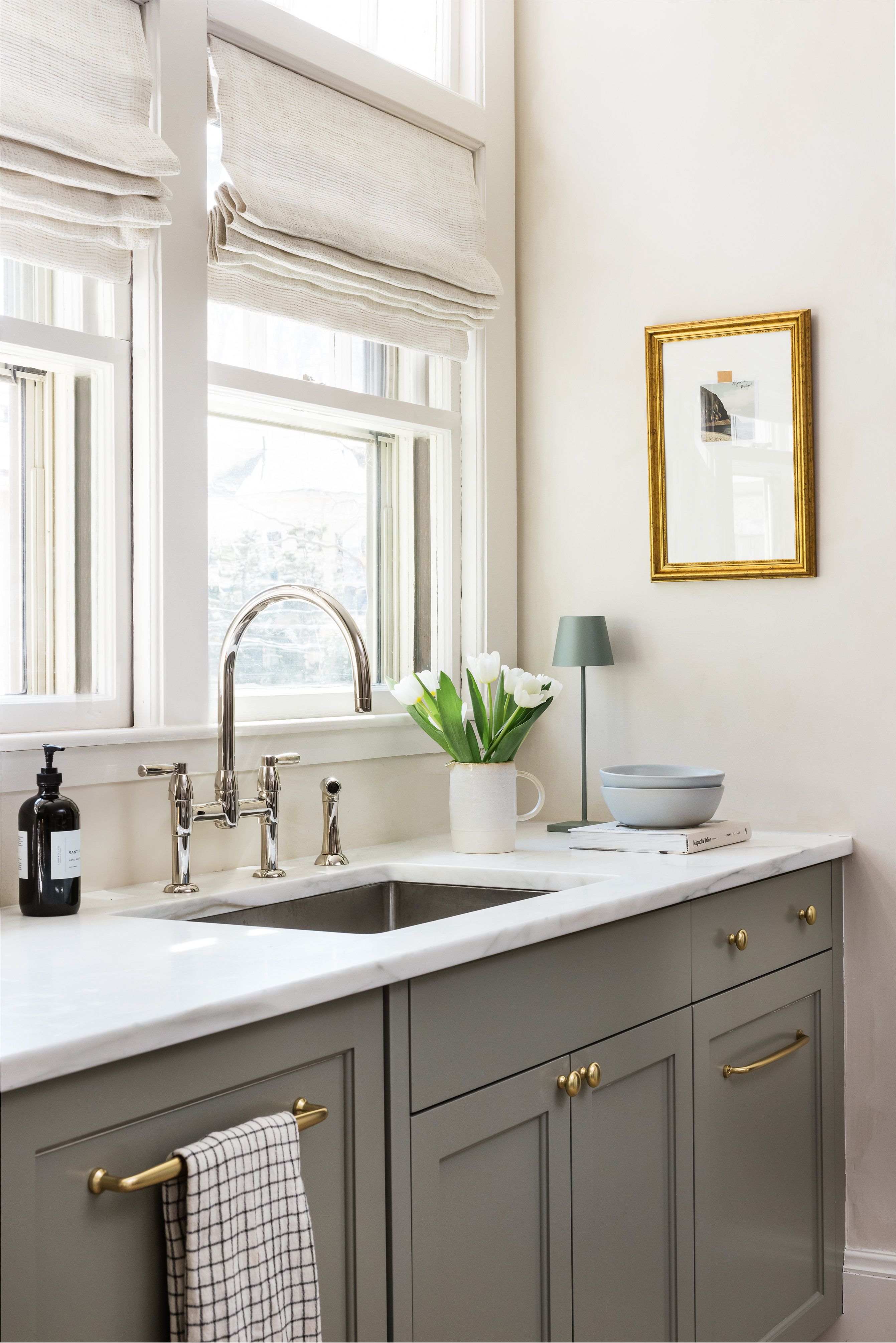 A kitchen sink area with an arched polished nickel faucet, white tulips in a pitcher, a gold-framed print, and marble countertops.