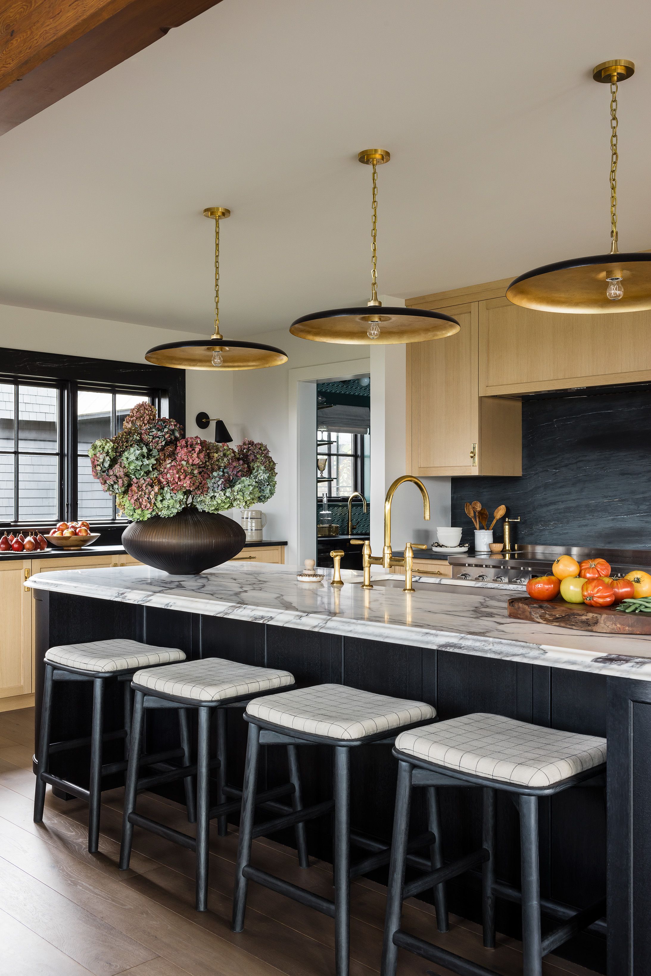 A kitchen island with a marble countertop sits beneath brass-and-black disc pendants, paired with black counter stools in checked fabric.