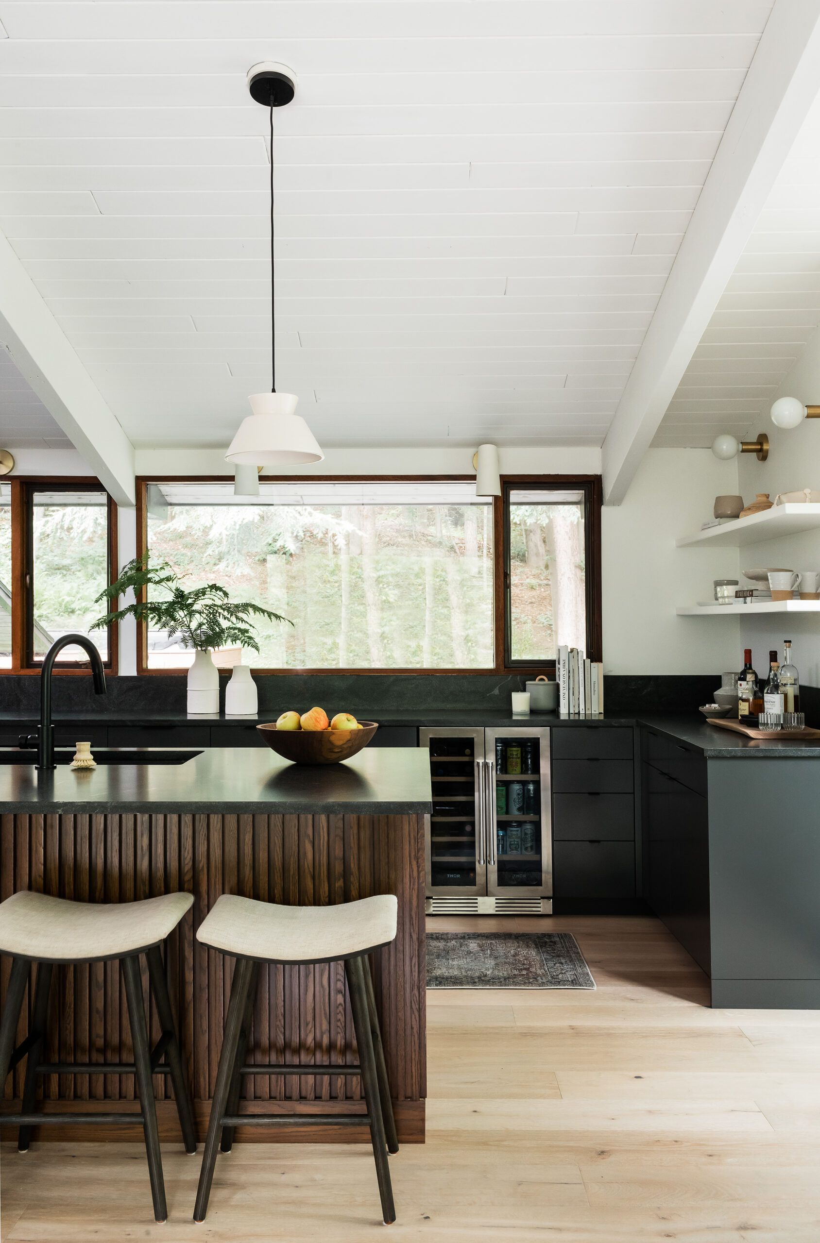 A kitchen with black cabinetry, a walnut slat-front island, dark stone countertops, and open shelving with ceramics.