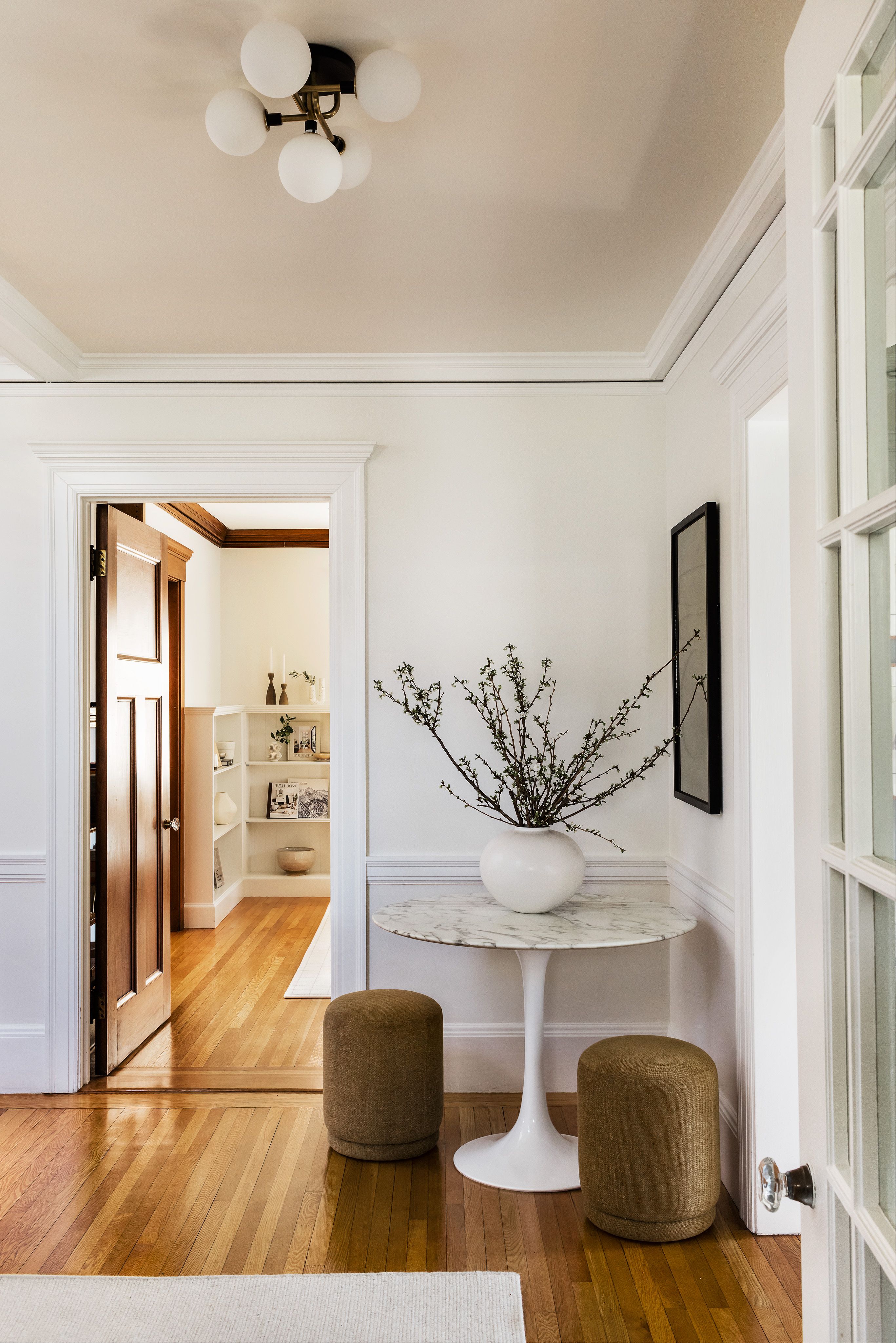 An entryway with a round marble table, branches in a white vase, olive green poufs, and a brass globe ceiling fixture.