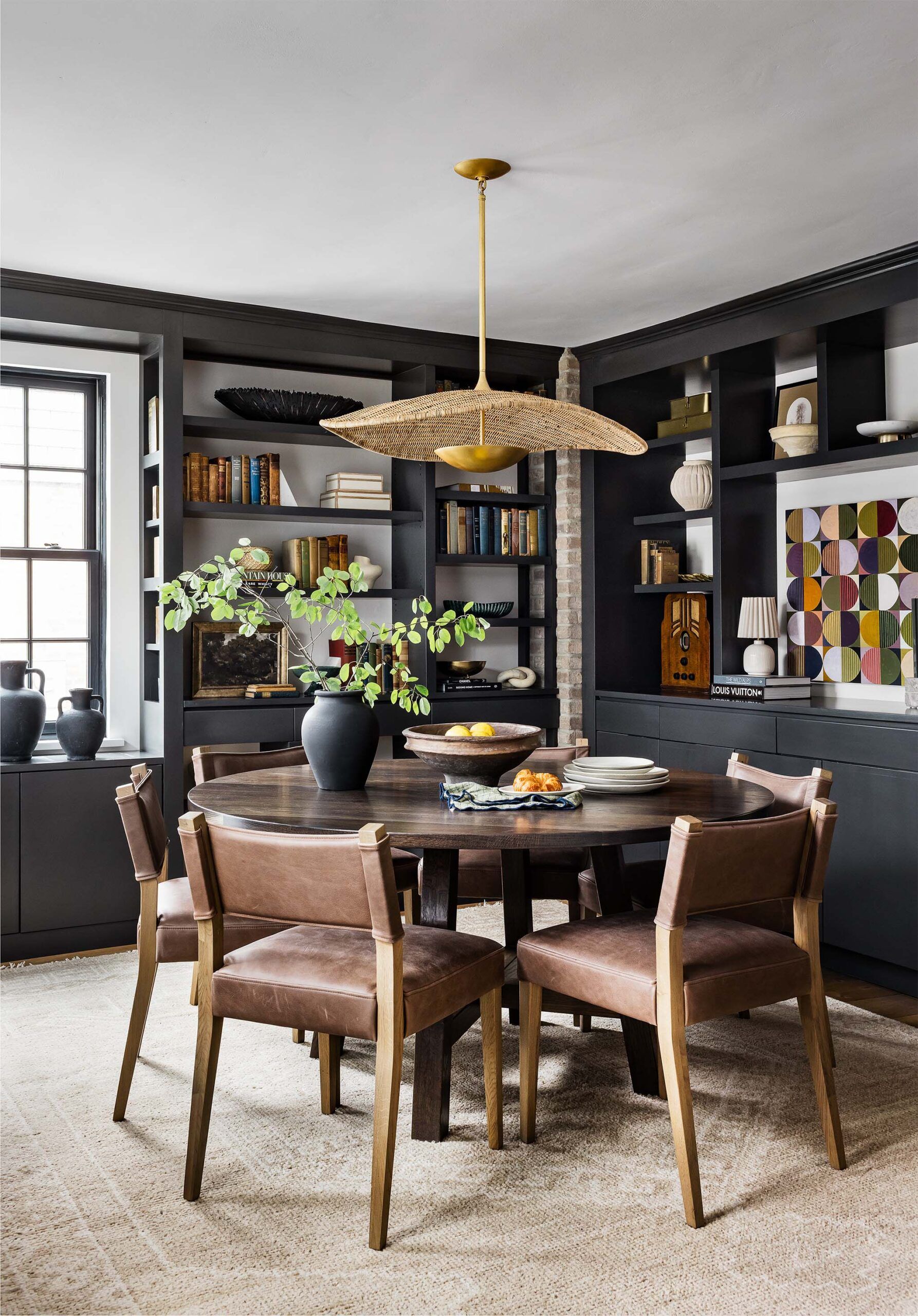 Dining room with a round dark wood table, leather dining chairs, built-in black bookshelves, and a woven rattan pendant light.