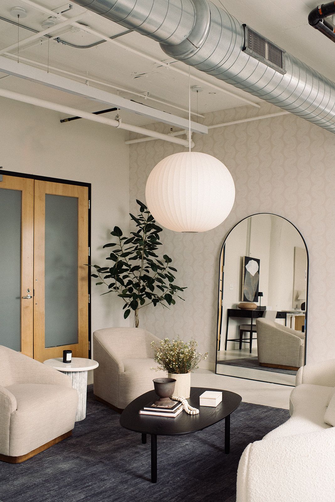 Lounge area with a round pendant light, an arched mirror, wood louvered doors, beige armchairs, and a charcoal rug.