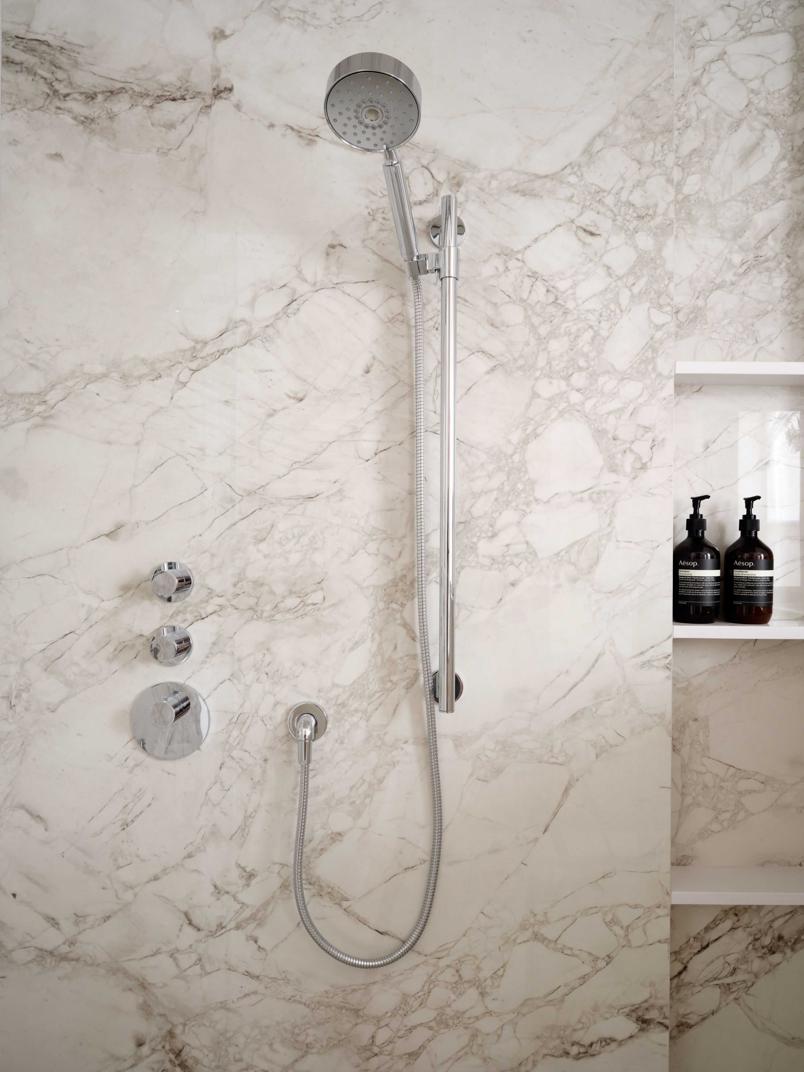 A marble-clad shower with a chrome handheld sprayer on a slide bar and a built-in niche holding bath product bottles.