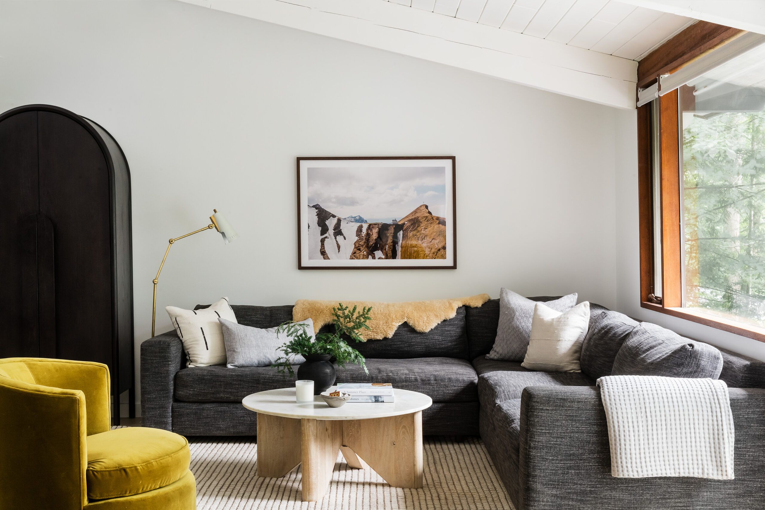 A living room with a charcoal sectional sofa, mustard velvet swivel chair, marble-top coffee table, and a framed mountain photograph.