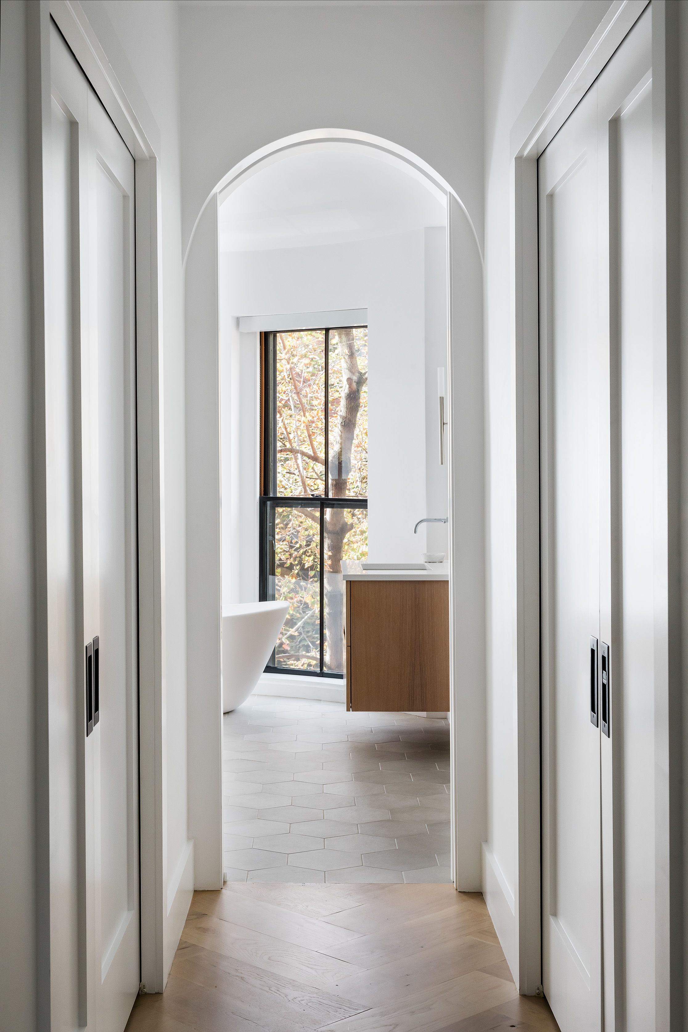 A hallway framed by an arched doorway and pocket doors leads to a bathroom with a freestanding tub and oak vanity.