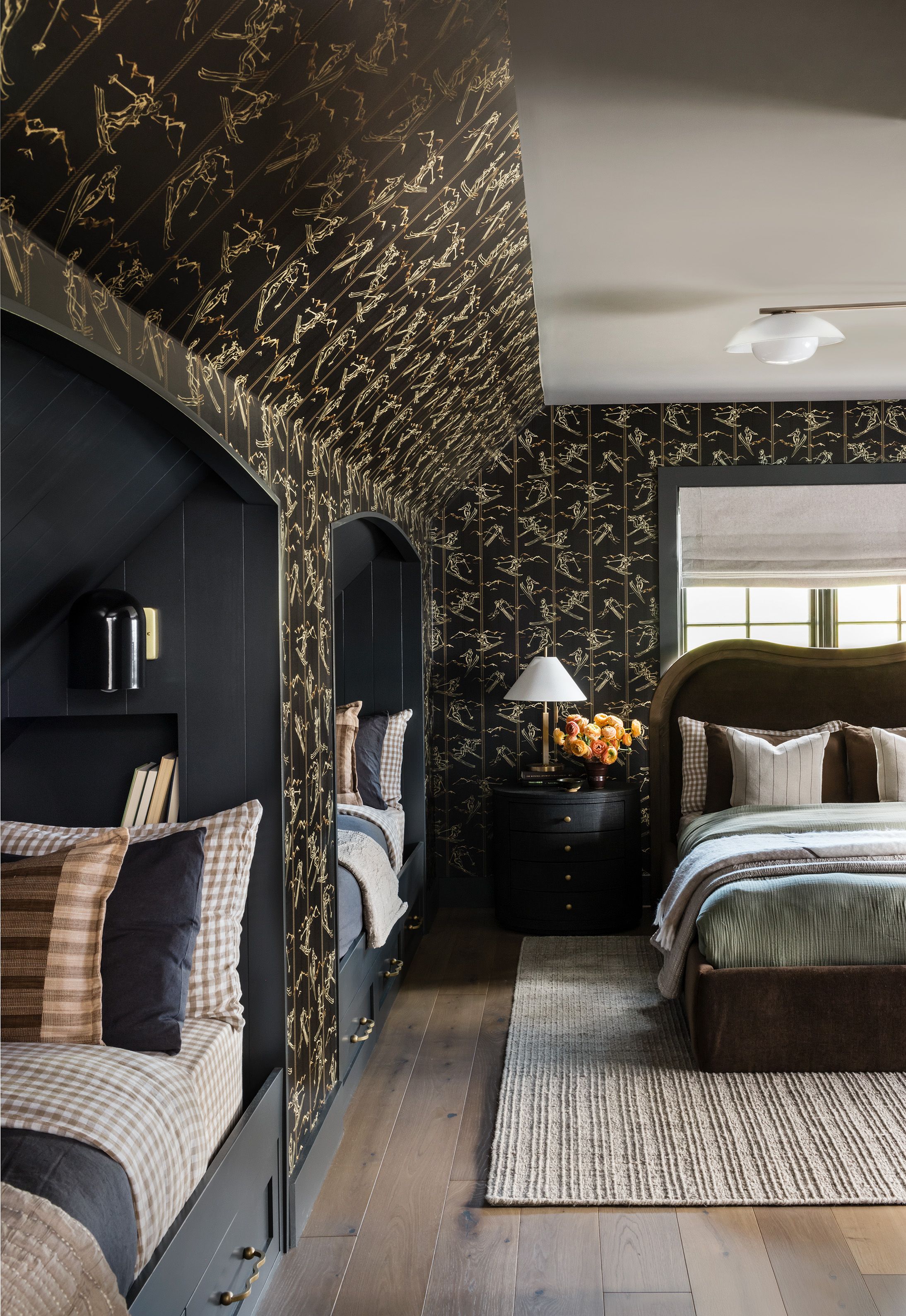 A dark attic bunk room with gold ski-print wallpaper has twin beds with gingham pillows beside a round black nightstand and brass lamp.