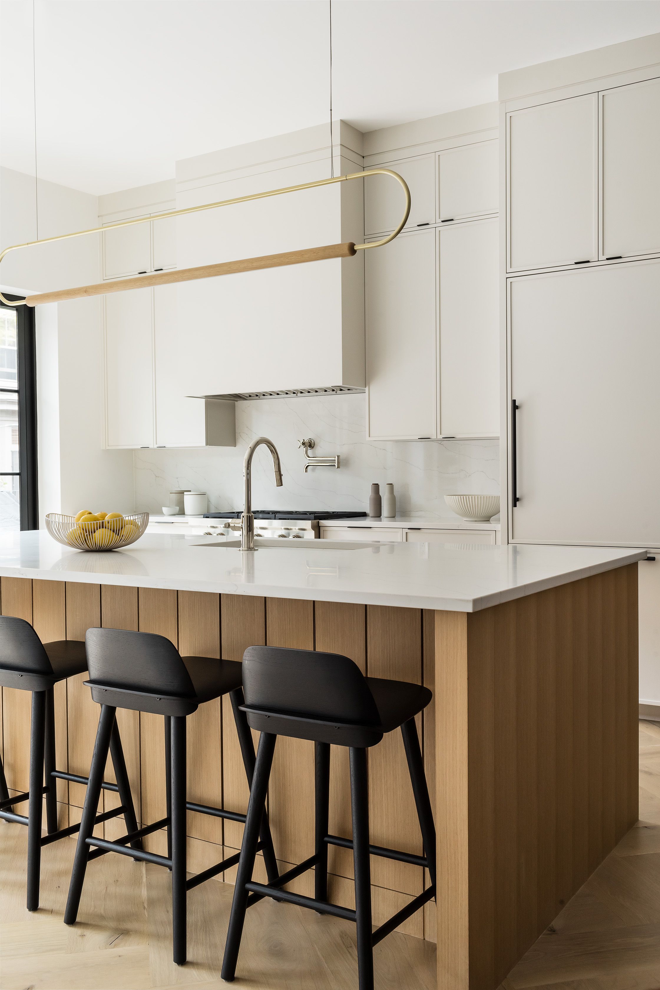 Kitchen island topped in white marble with black barstools, a wire fruit basket, and a curved brass pendant light overhead.