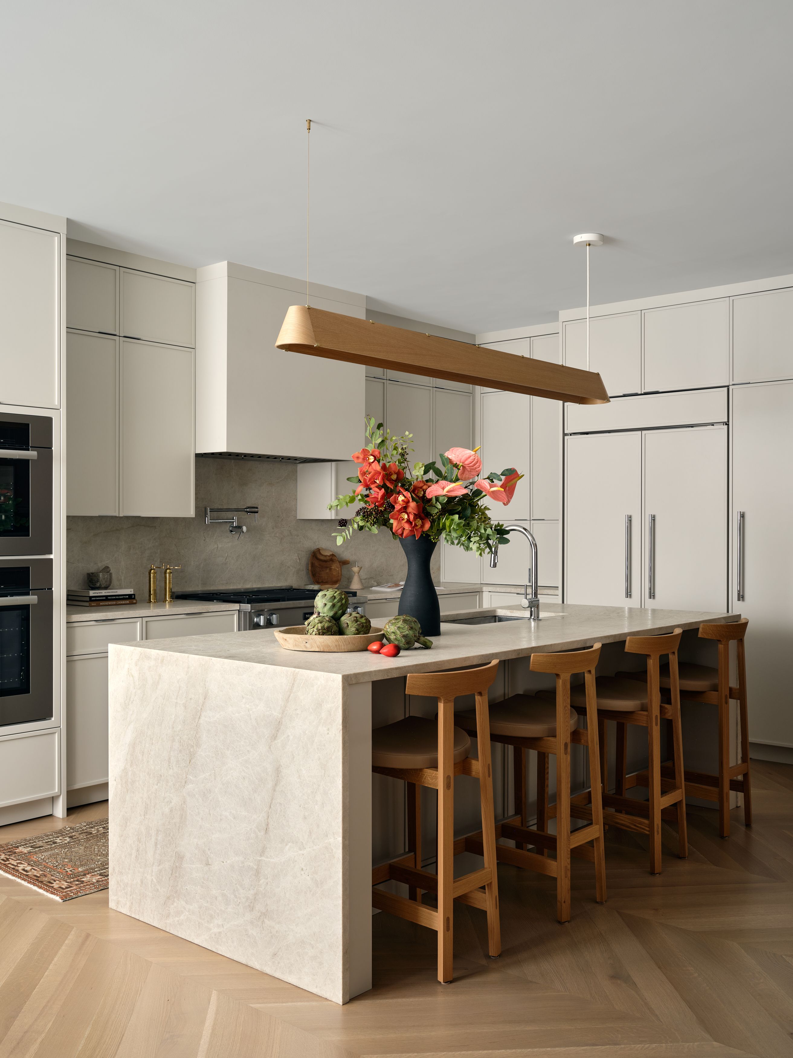 A kitchen island with a wood pendant light, marble waterfall counter, an orange floral arrangement, and wood bar stools.