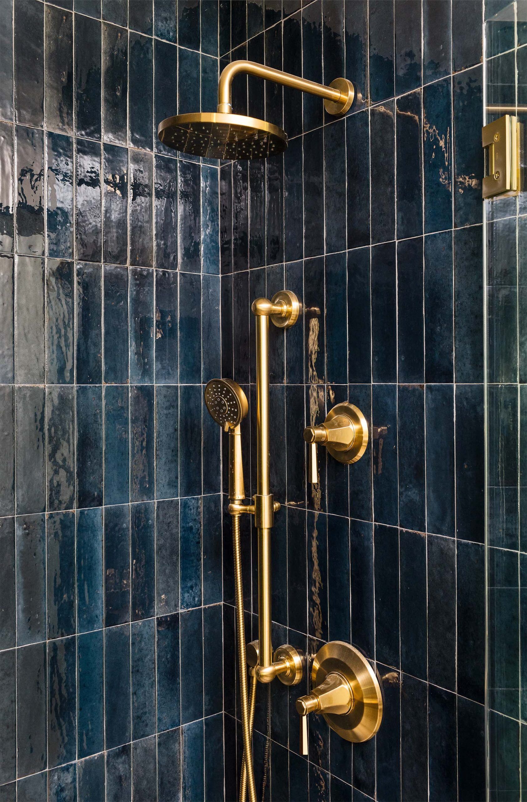 Shower detail with glossy dark blue vertical tile and brass fixtures, including a rain shower head and a handheld sprayer.