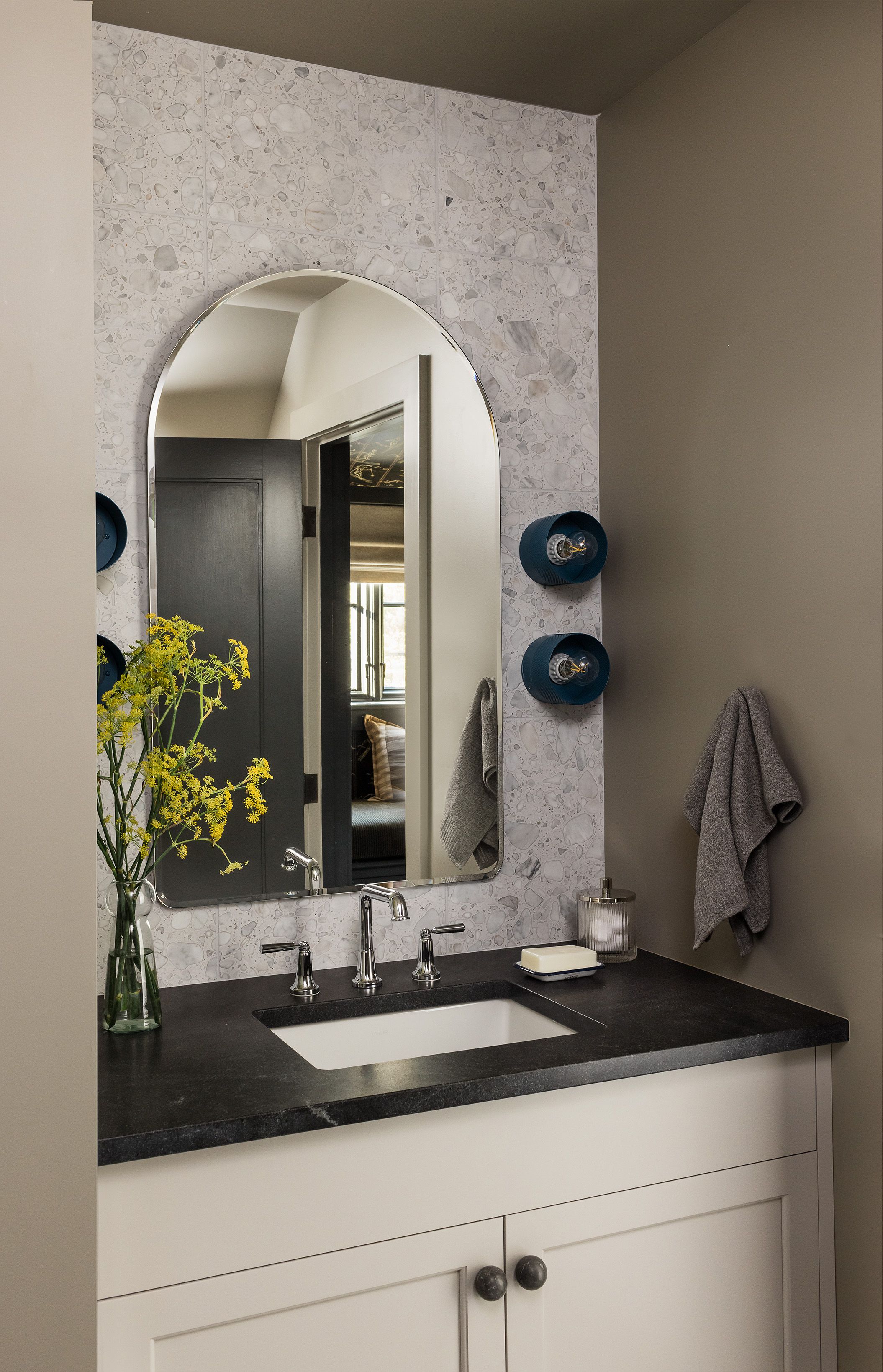 A bathroom with terrazzo-tile walls pairs an arched mirror and navy barrel sconces above a black stone vanity counter with an undermount sink.