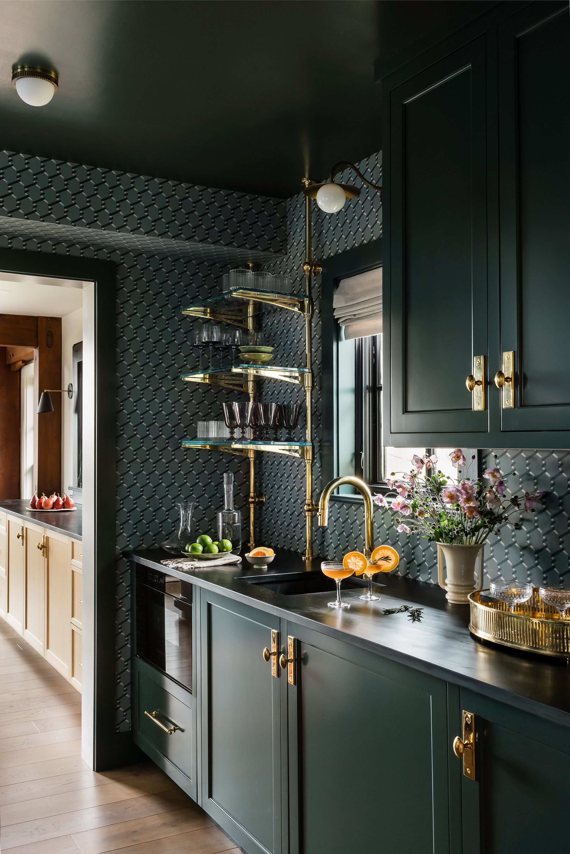 A butler's pantry in dark green cabinetry pairs patterned wallpaper with brass ladder shelving for glassware and a black stone counter.