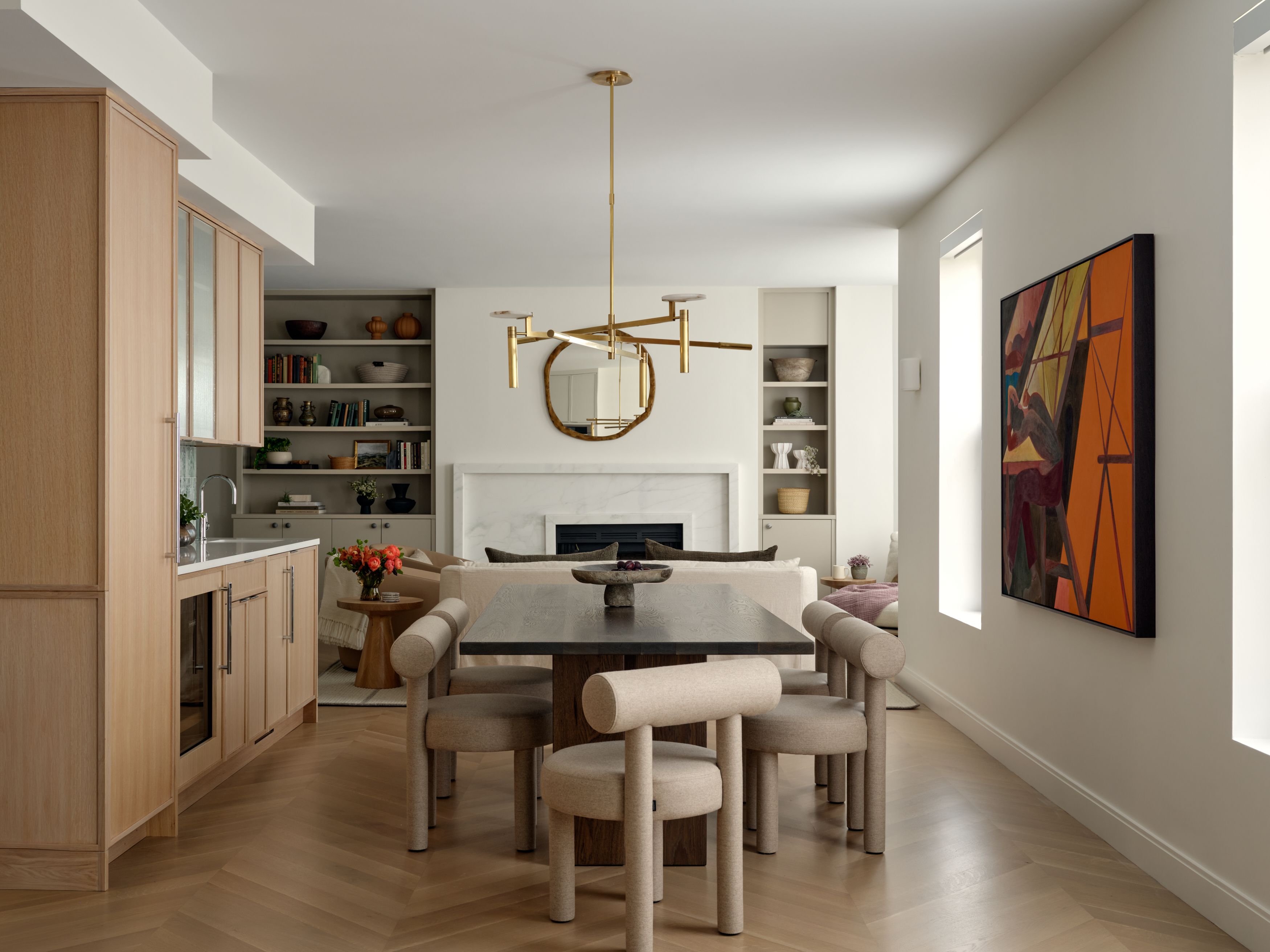 A dining area with a brass sculptural chandelier, a round mirror over a marble fireplace, and an orange abstract painting.