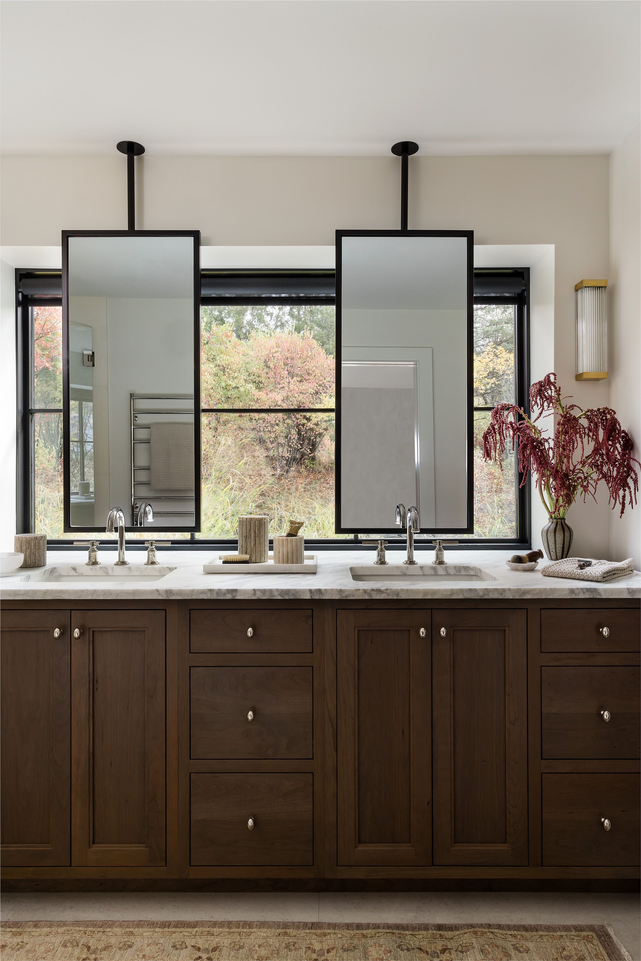 A primary bathroom's dark wood double vanity has a white marble counter under two ceiling-hung rectangular mirrors facing a woodland window.