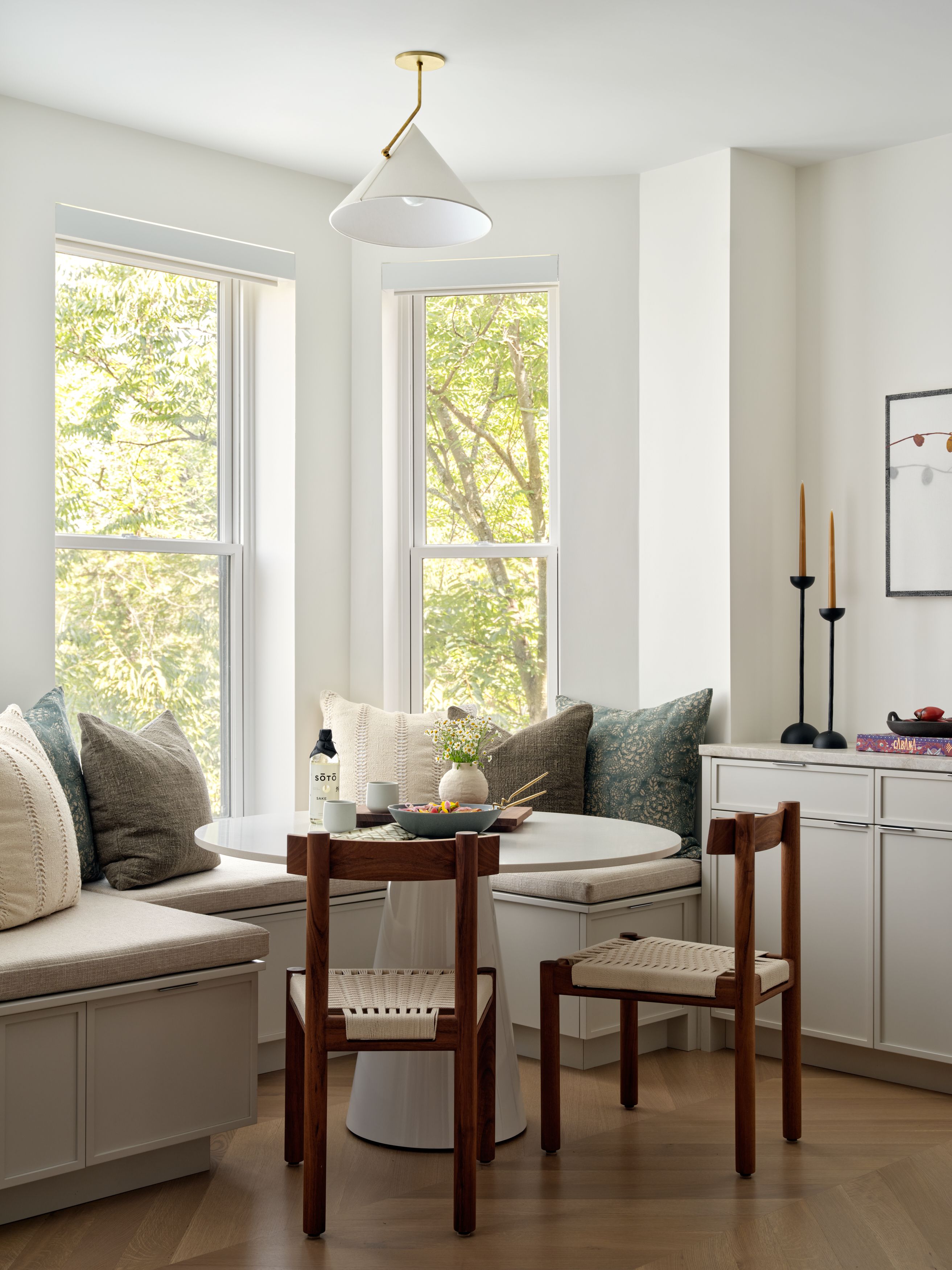 A bay-window breakfast nook with a cushioned built-in banquette, woven-seat wood chairs, and a round pedestal table.