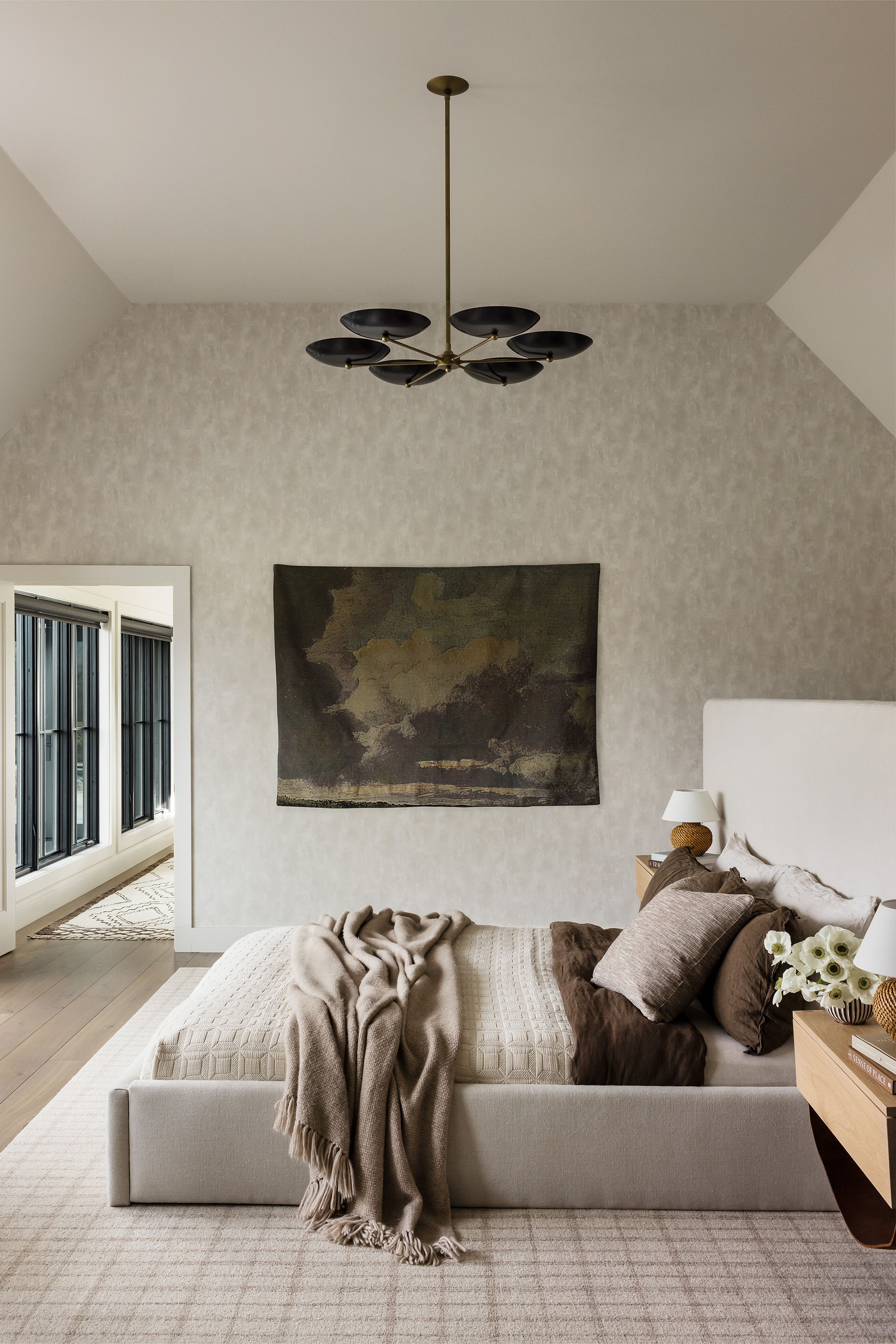 A primary bedroom bed with cream and brown linens sits beneath a moody cloud tapestry and a black-and-brass sculptural chandelier.
