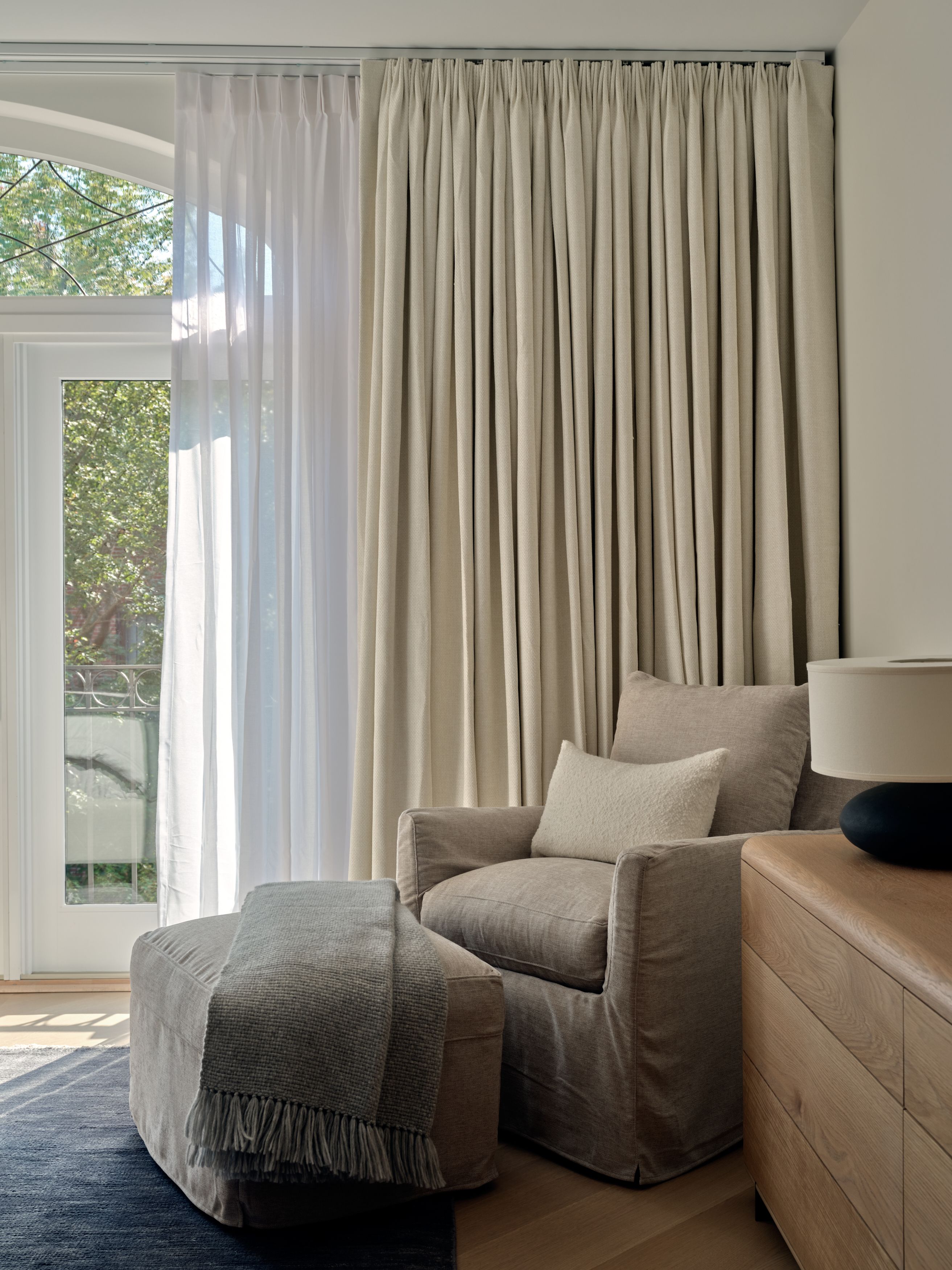 A bedroom seating corner with sheer and linen drapery, an upholstered swivel chair, a bouclé ottoman, and a wood dresser.