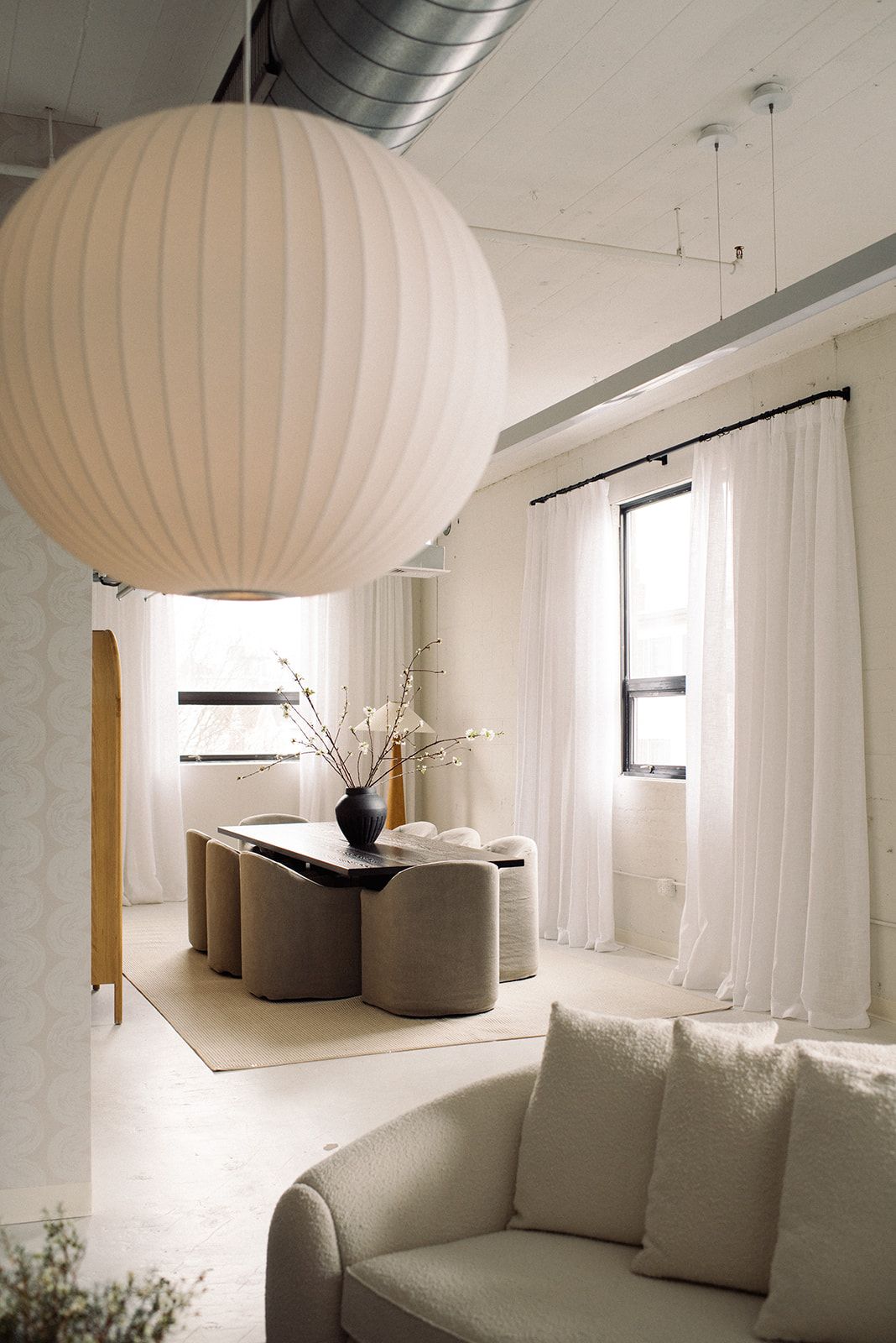 A globe paper lantern pendant hangs over a dining table with curved taupe chairs, a black vase of blossom branches, and a boucle sofa.