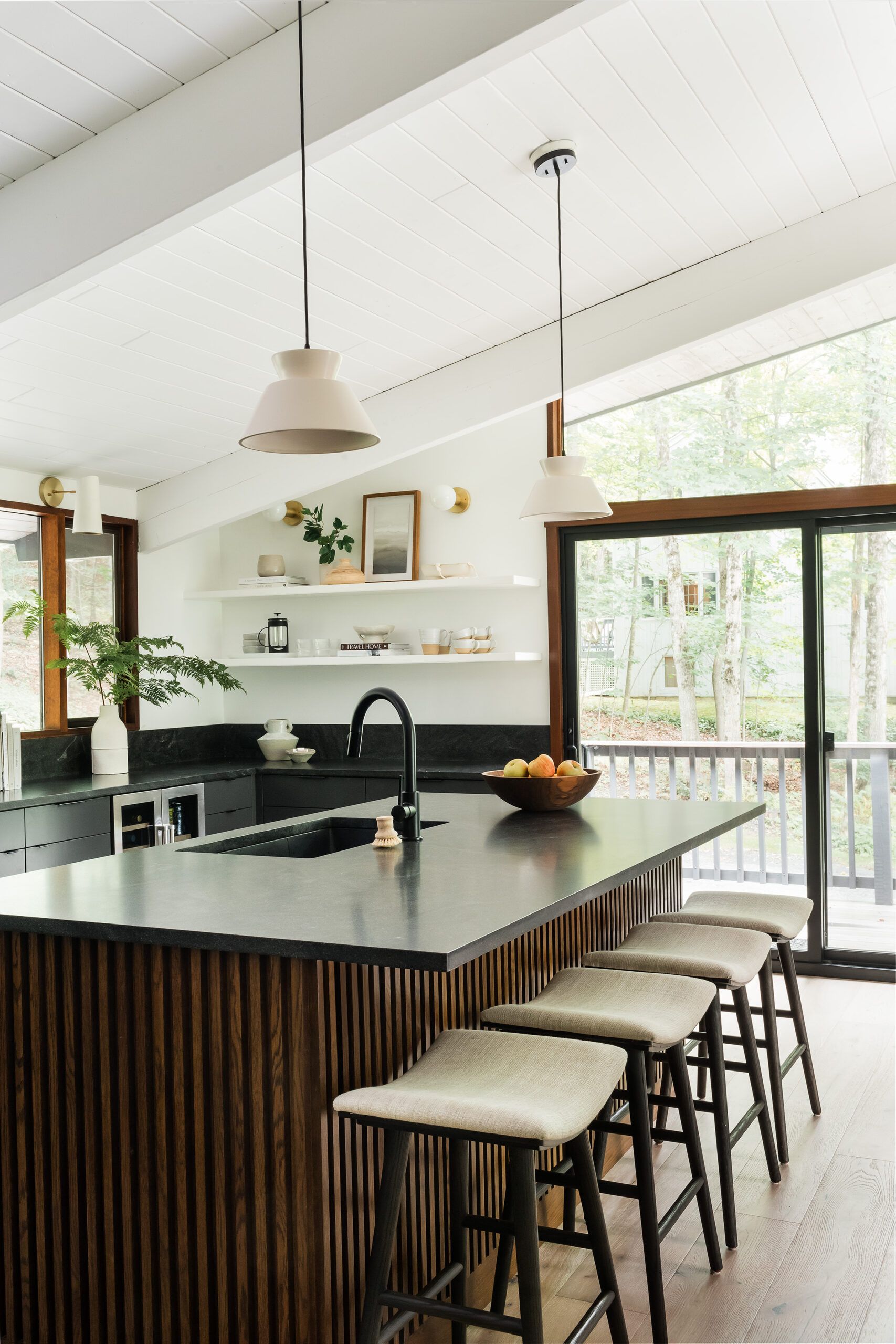 A kitchen island with a dark stone top, walnut slat base, wood bar stools, and two white pendant lights over a view of the yard.