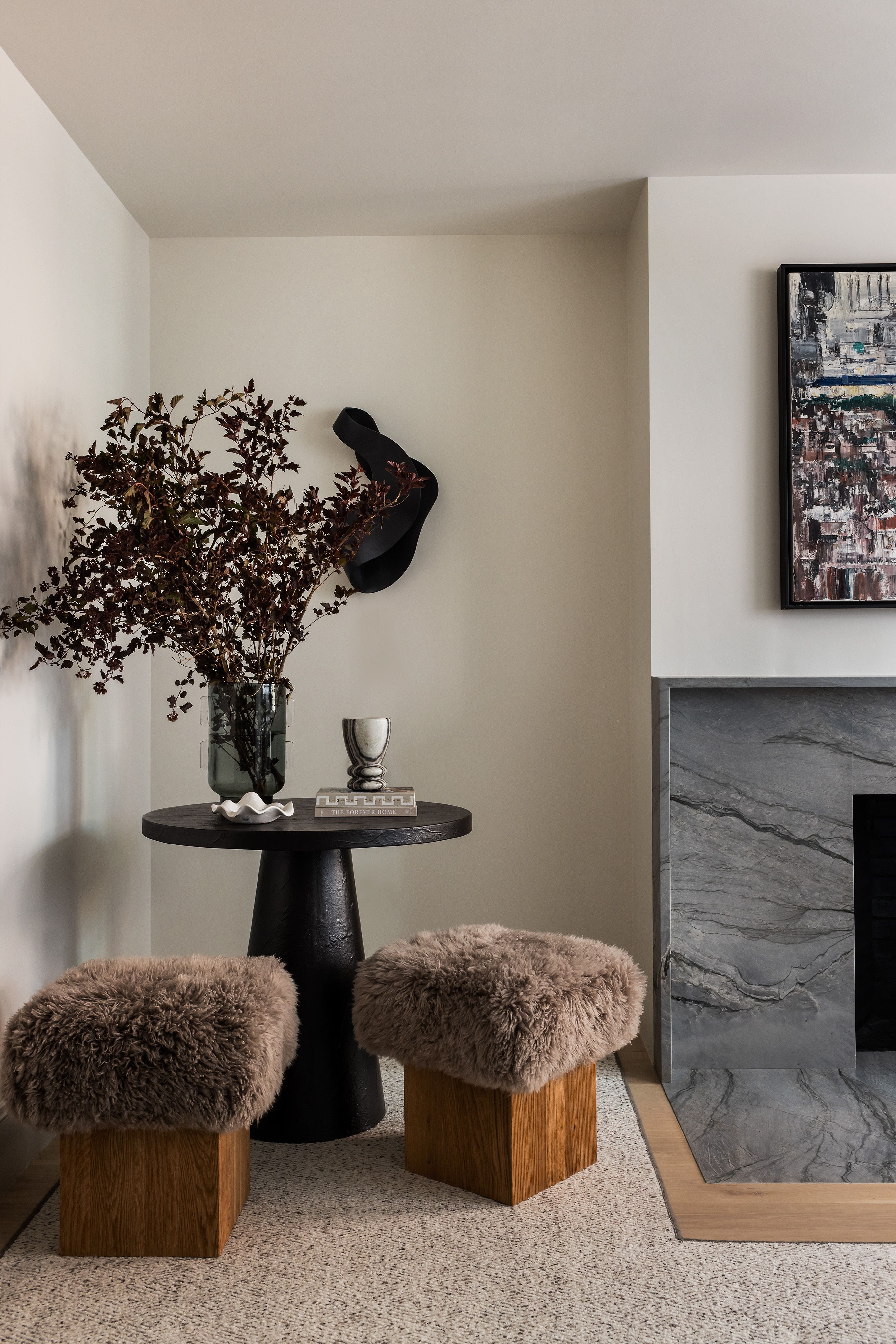 A living room corner with a black pedestal table holding dried branches, a black wall sculpture, and gray sheepskin stools.