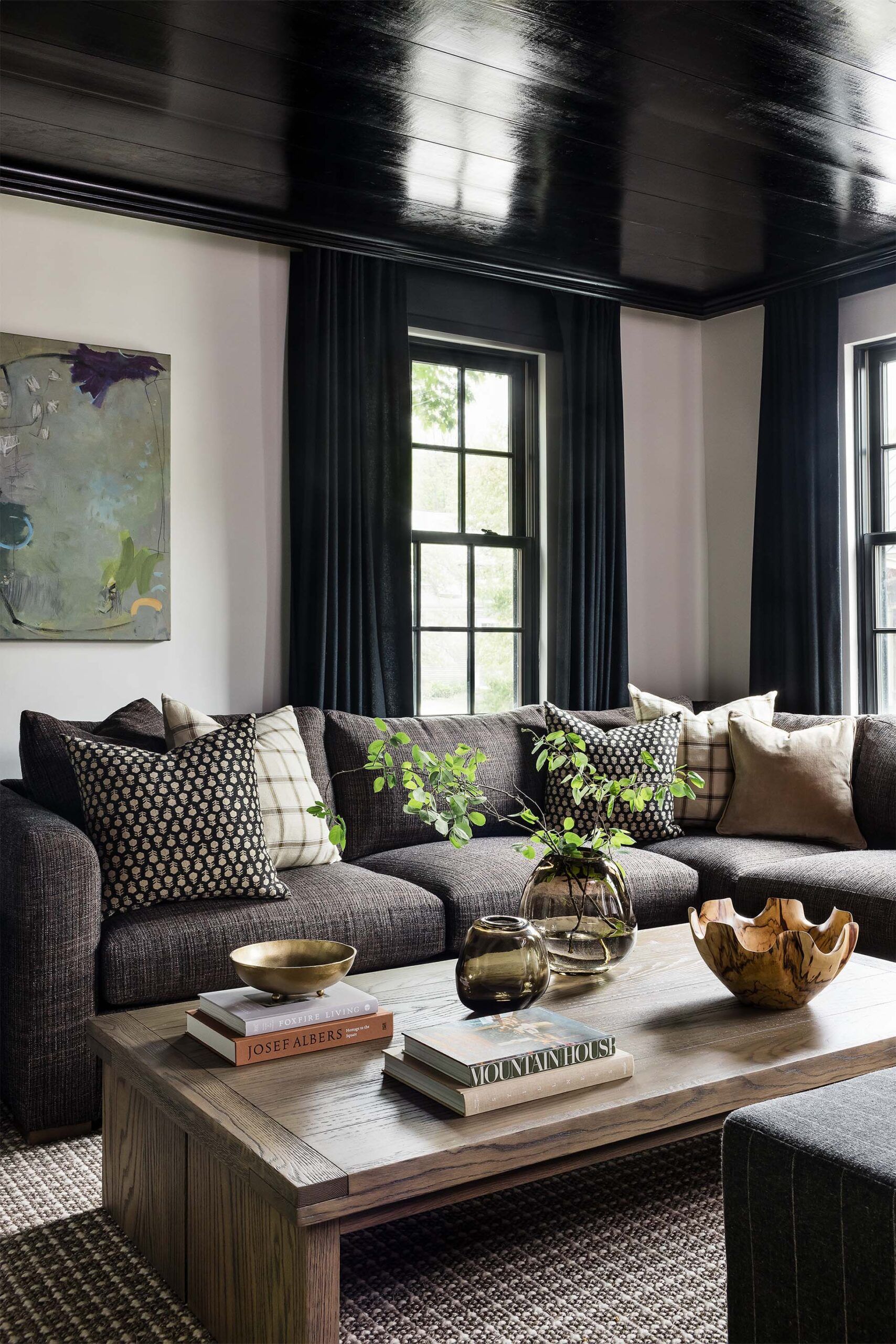 Living room with a charcoal sectional sofa, patterned throw pillows, black-trimmed windows, and a wood coffee table with vases.