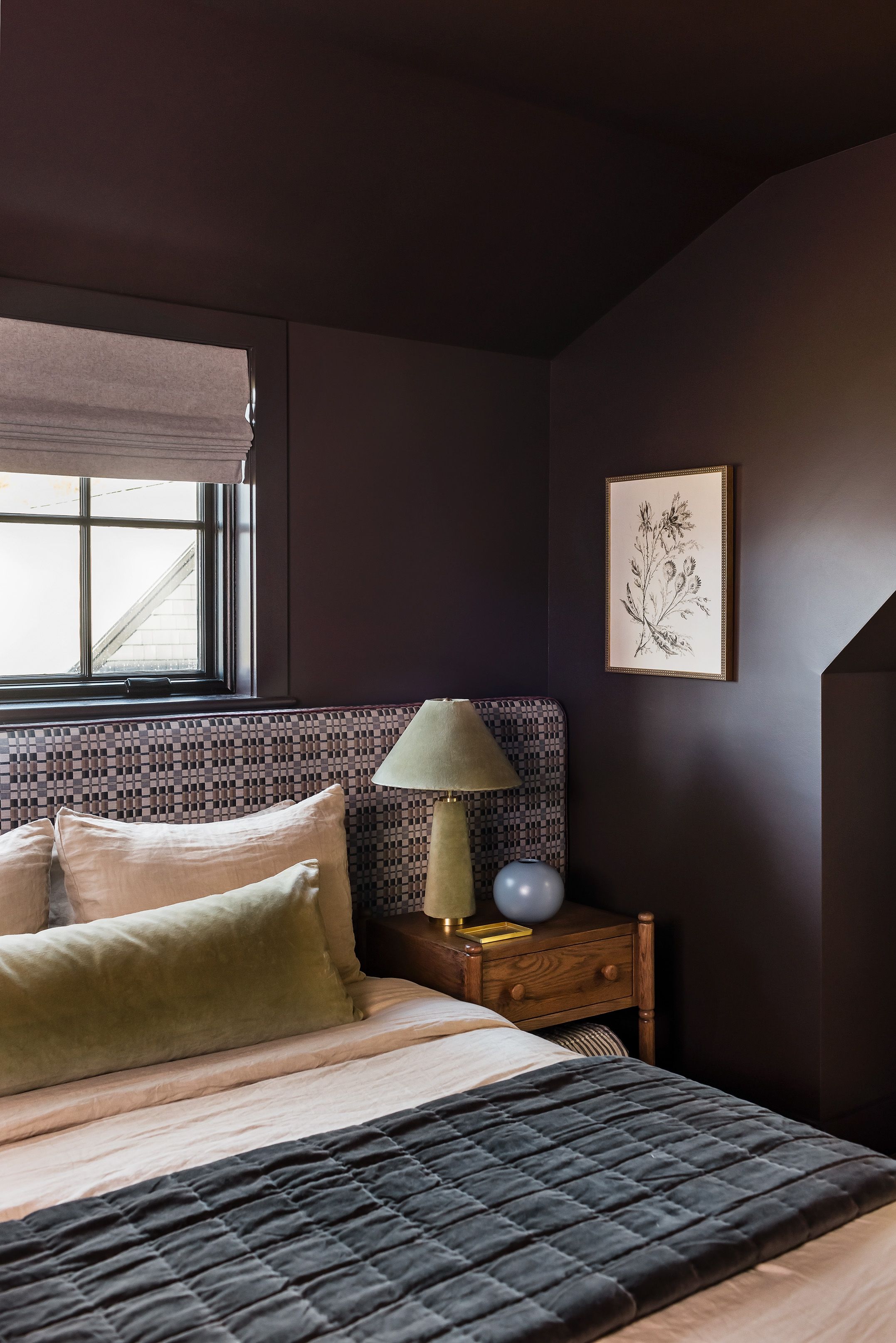 A dark plum bedroom's wood nightstand holds a green velvet lamp and a round blue vase beside a framed botanical illustration.