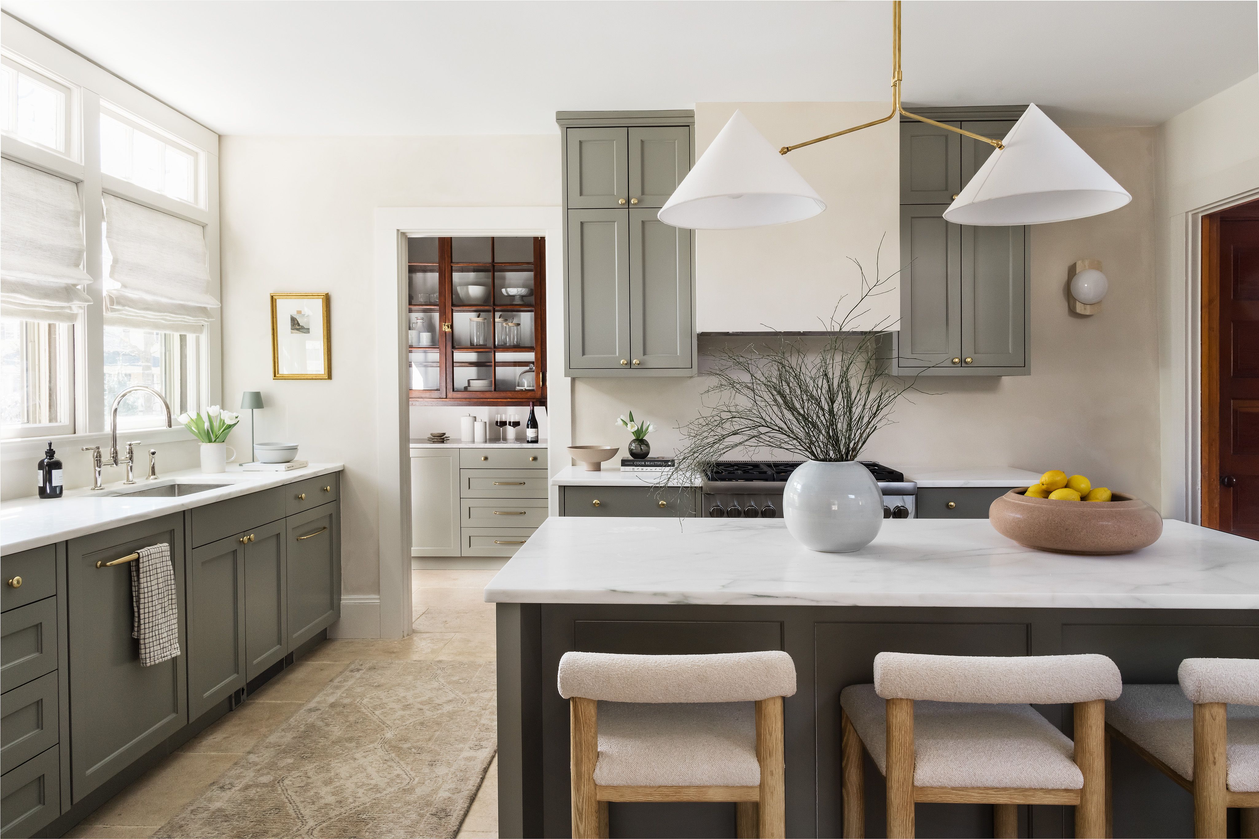 A kitchen island with cone-shaped pendant lights, sage green cabinetry, a bowl of lemons, and an abstract painting on the wall.