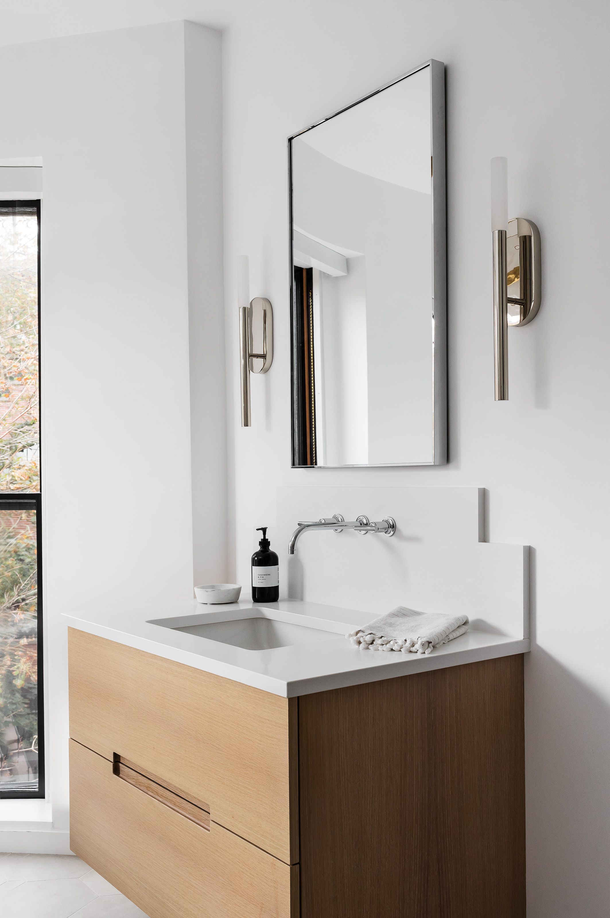 A bathroom vanity with a frameless mirror, nickel wall sconces, and a two-tone oak cabinet beneath a white stone counter.