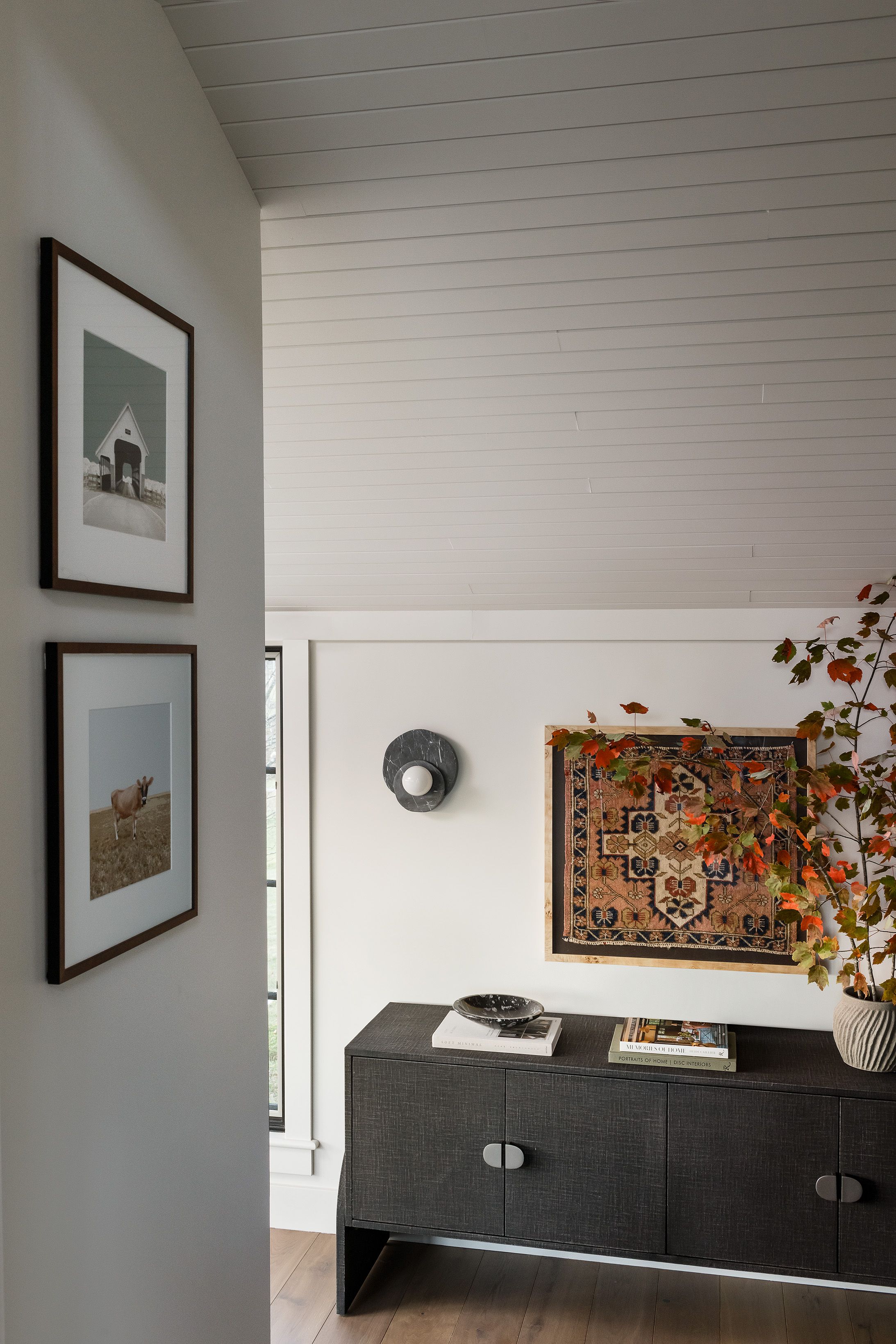 A hallway with framed farm photography leads to a dark woven credenza topped with a vintage rug tapestry and a ceramic vase of branches.