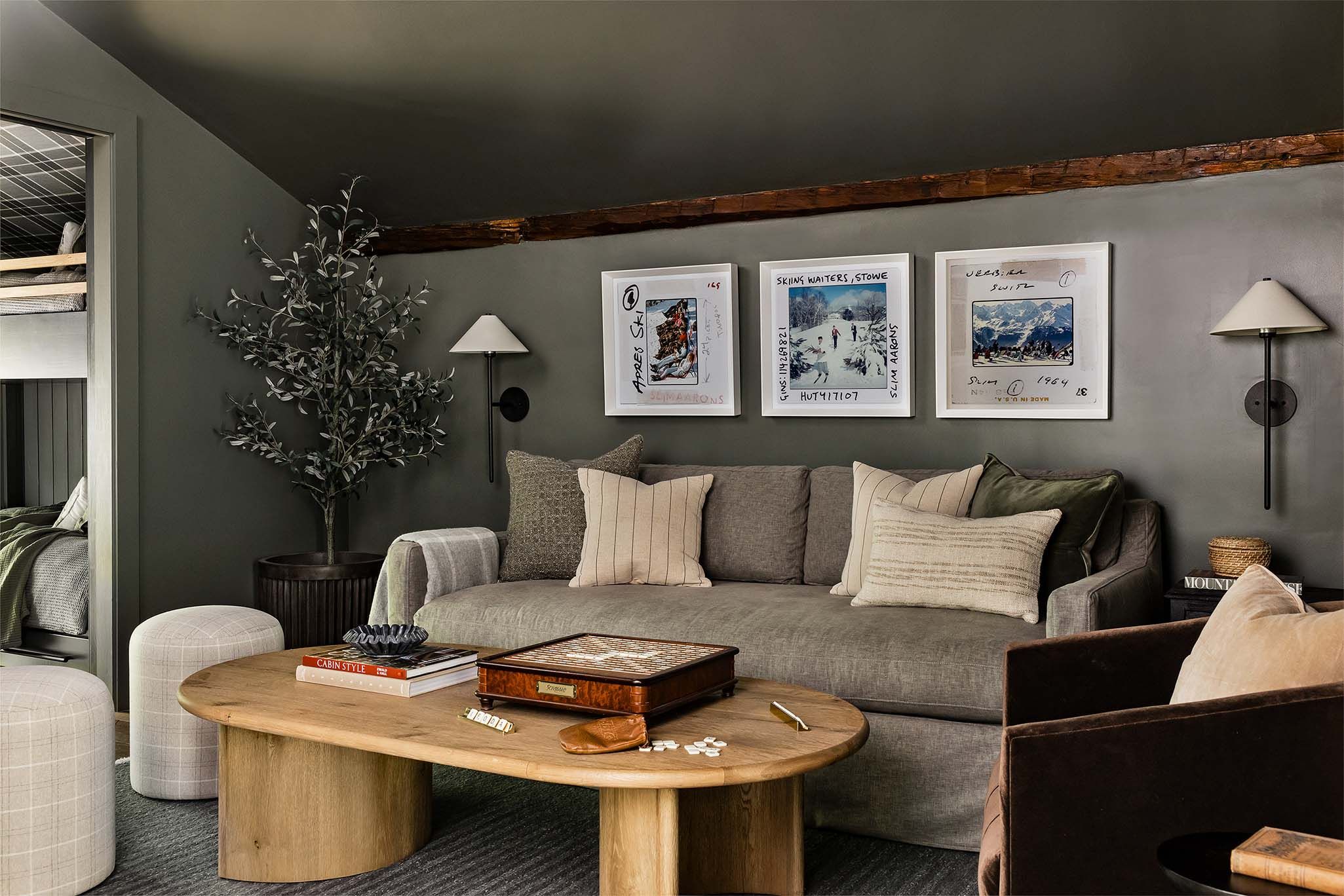 Den with olive-painted walls, a rustic wood beam, a gray sectional sofa, framed ski photos, and a round wood coffee table.