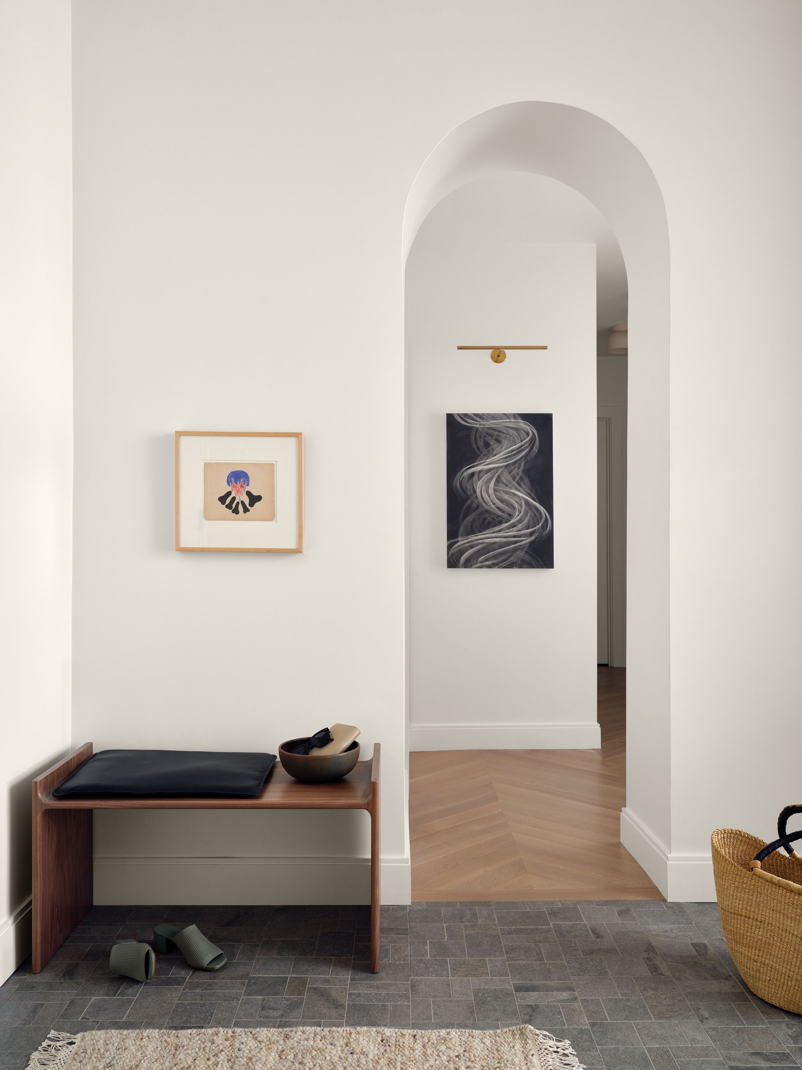 An entryway with a walnut bench, a woven basket, and an arched doorway revealing an abstract black-and-white painting beyond.