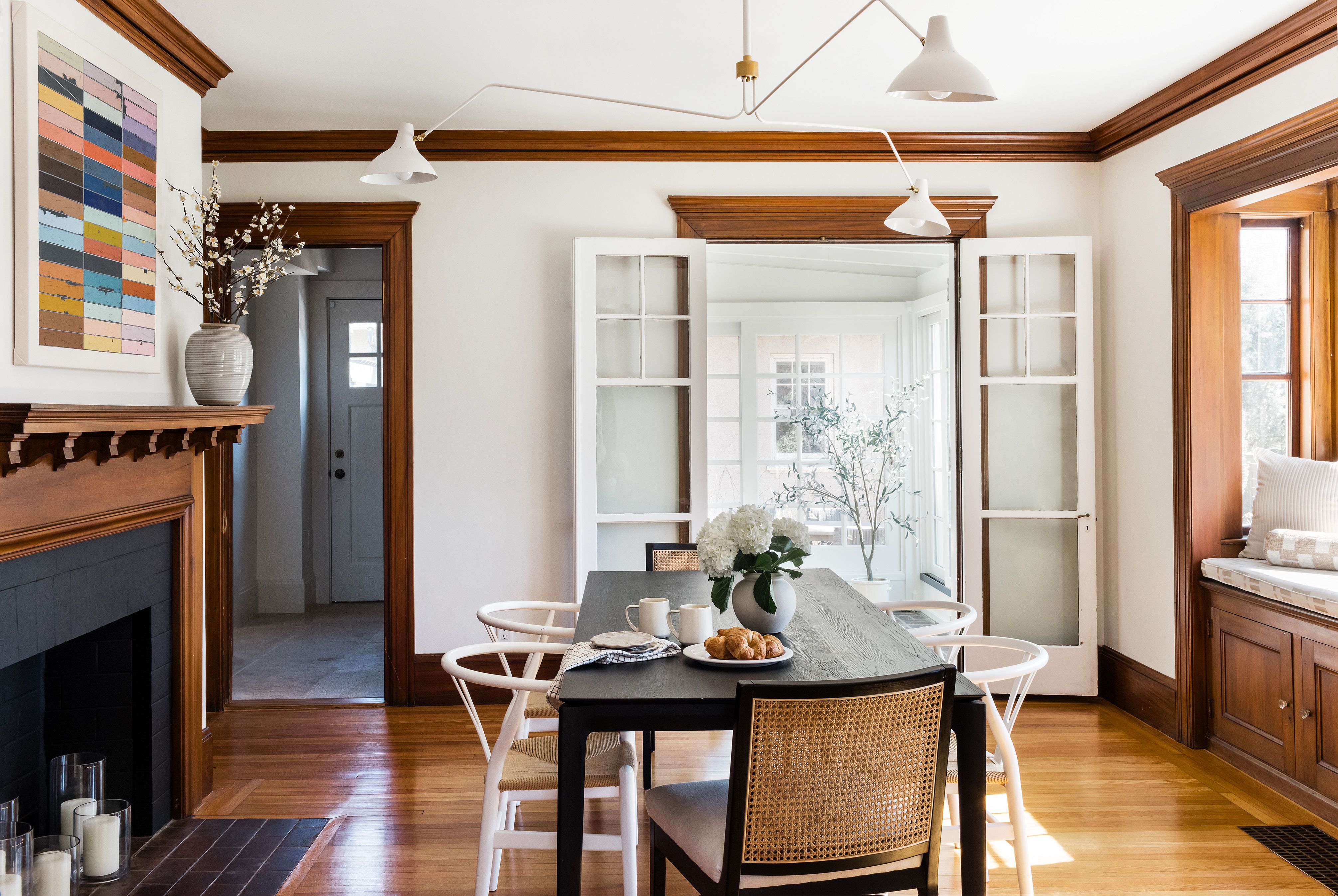 A dining room with a black table, a cane-back chair, a colorful abstract painting over the fireplace, and French doors open to a sunroom.