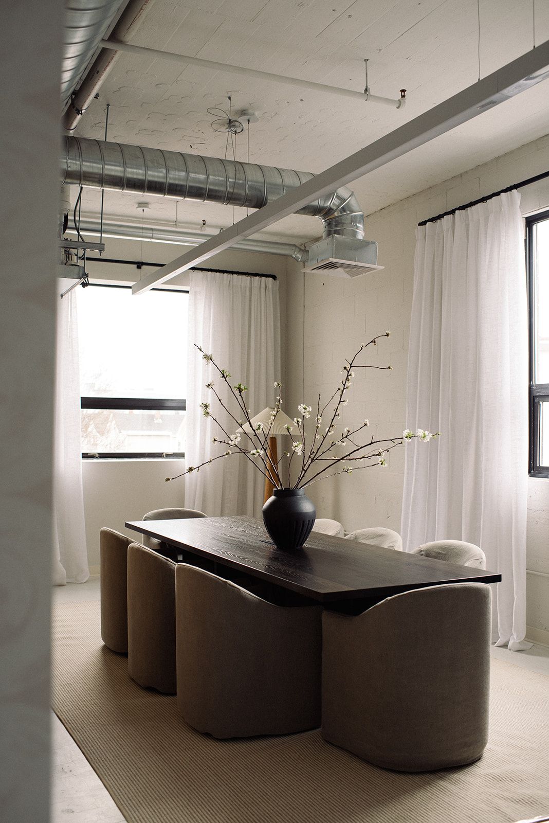A studio dining area with a dark wood table, cream bouclé chairs, flowering branches in a black vase, and floor-to-ceiling white curtains.
